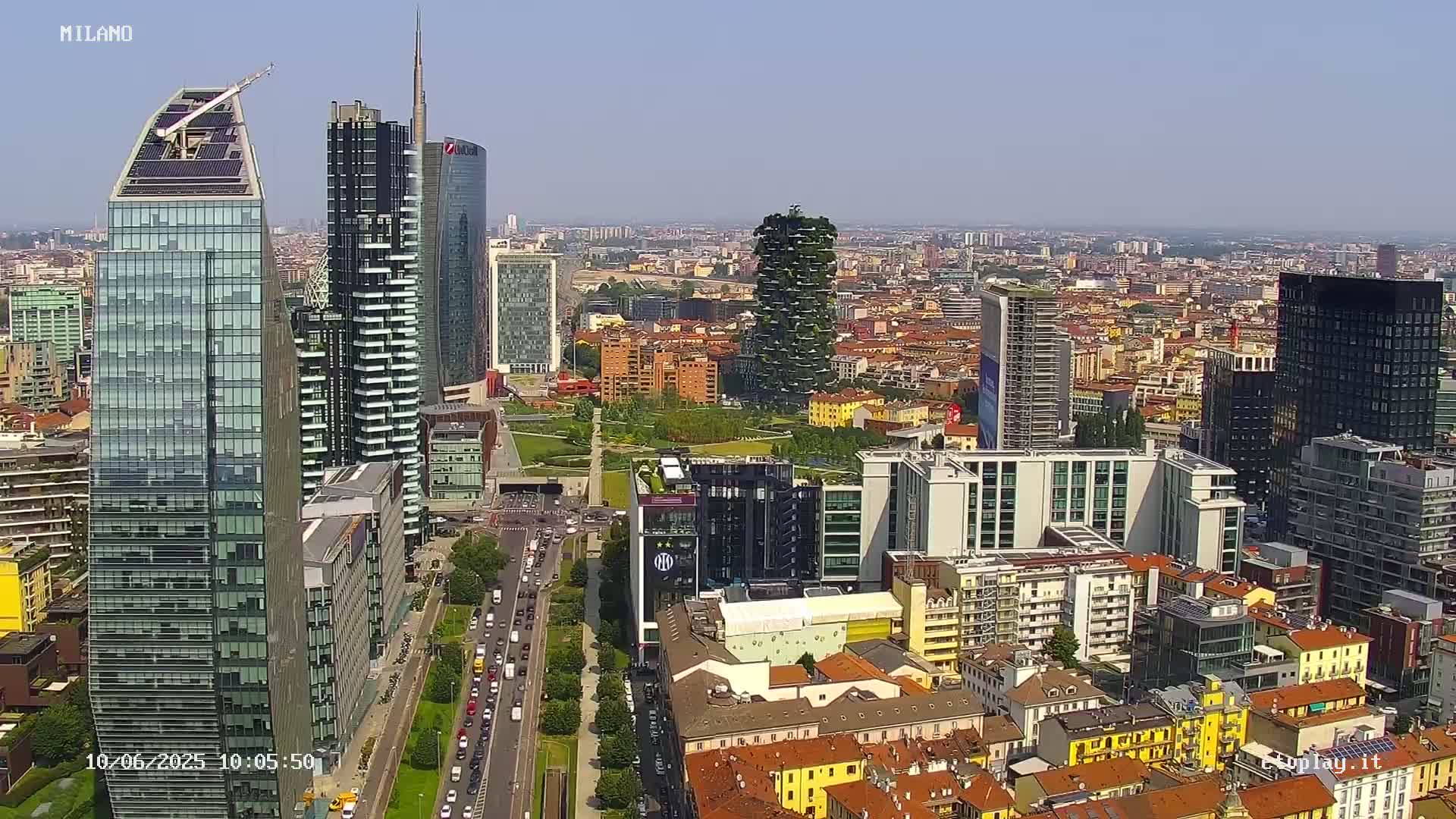 A high-angle, long shot of a city skyline on a clear, sunny day, showing numerous skyscrapers and surrounding buildings, with a roadway and park visible.