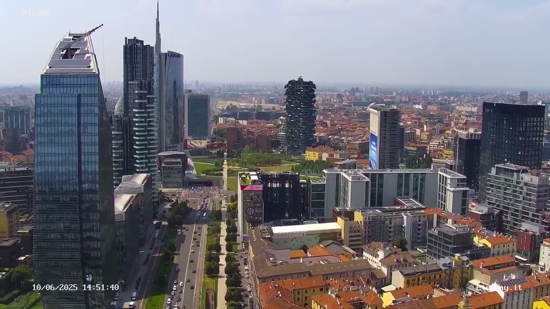 An aerial view of a city on a clear, sunny day, showing a mix of modern skyscrapers and older, low-rise buildings, with a park visible in the center.