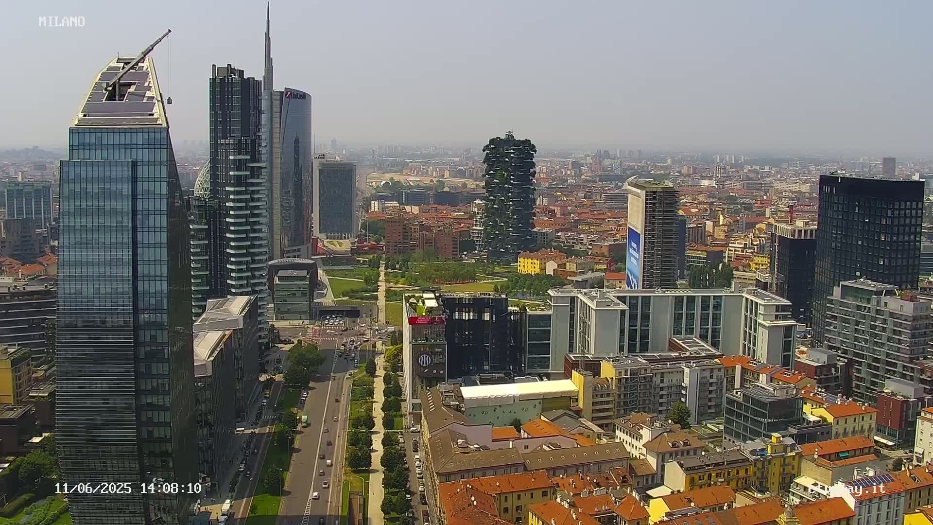 A hazy, sunny day view of a dense cityscape with numerous skyscrapers, including a tall, vertically-oriented green building, and a network of streets and buildings.