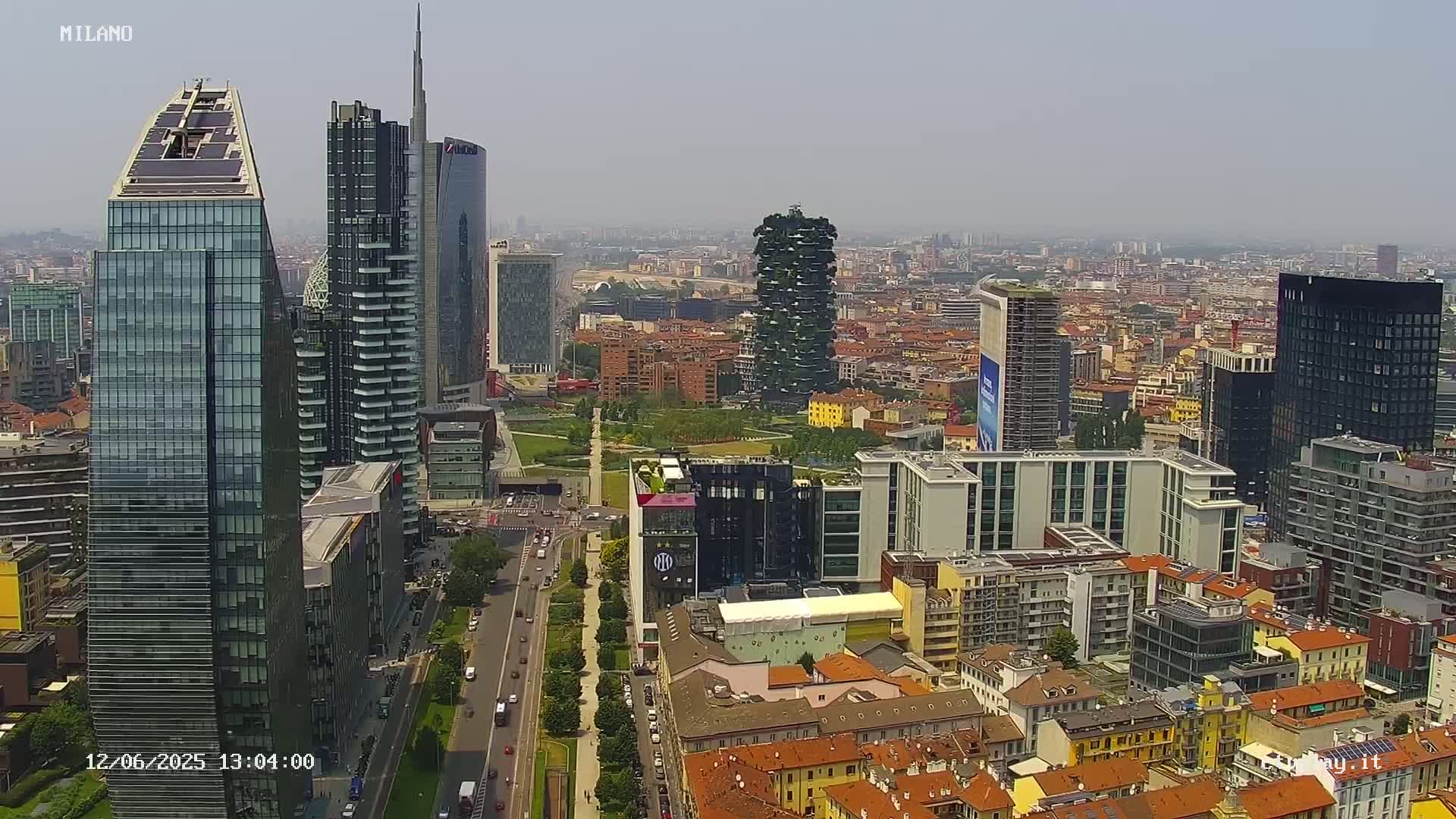 A hazy, sunny day view of a dense cityscape with numerous skyscrapers and modern buildings, including a tall, vegetated tower, and a wide street lined with trees.
