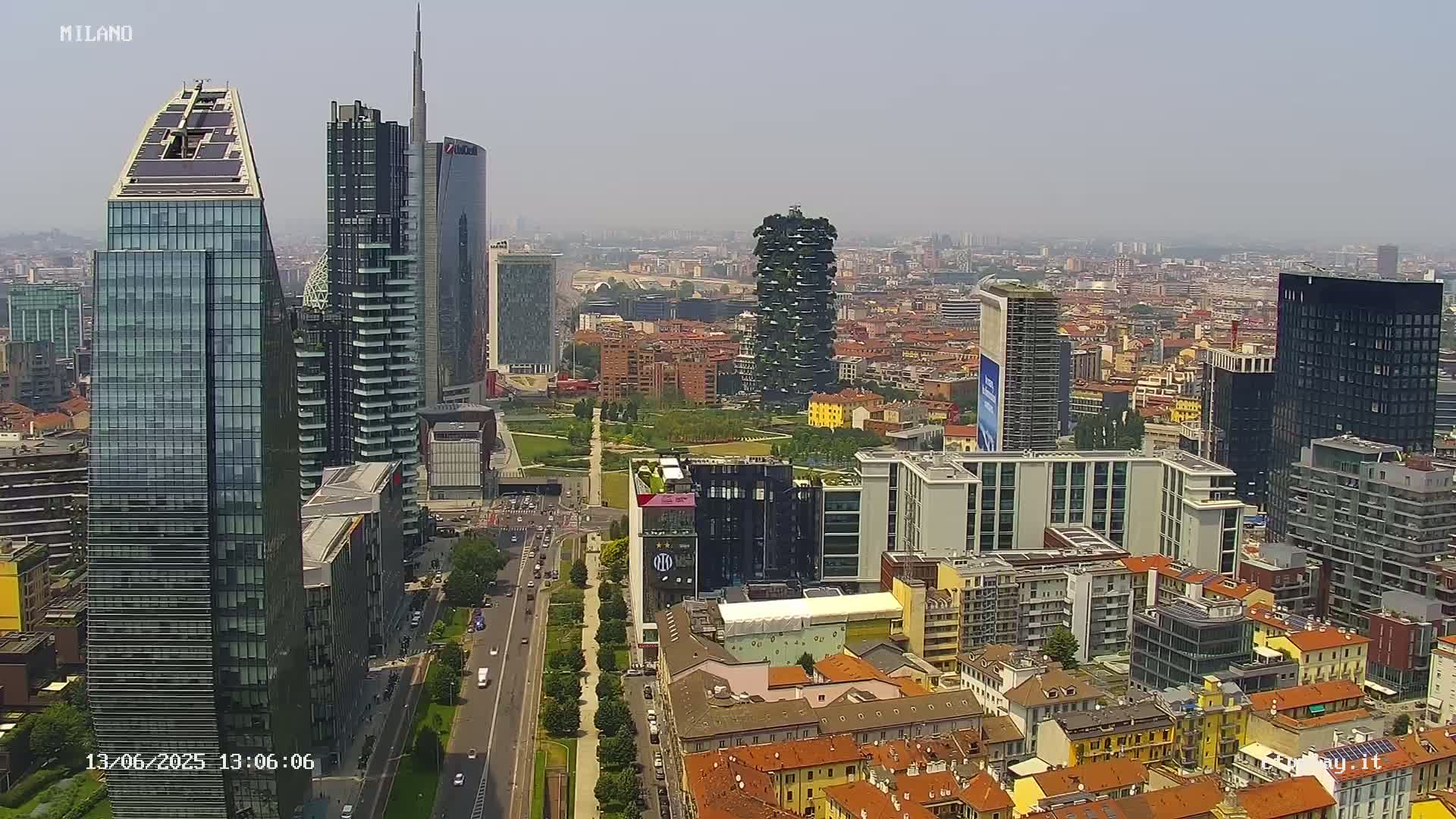 A high-angle, long shot of a city skyline on a hazy day, featuring numerous skyscrapers and buildings of varying heights and architectural styles, including a uniquely designed vertical forest building, with a wide street and a green park visible in the foreground.