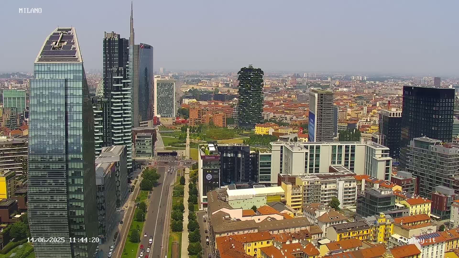 A high-angle, long shot of a city skyline on a clear, sunny day, showing numerous modern skyscrapers and surrounding buildings, interspersed with green spaces and tree-lined streets.