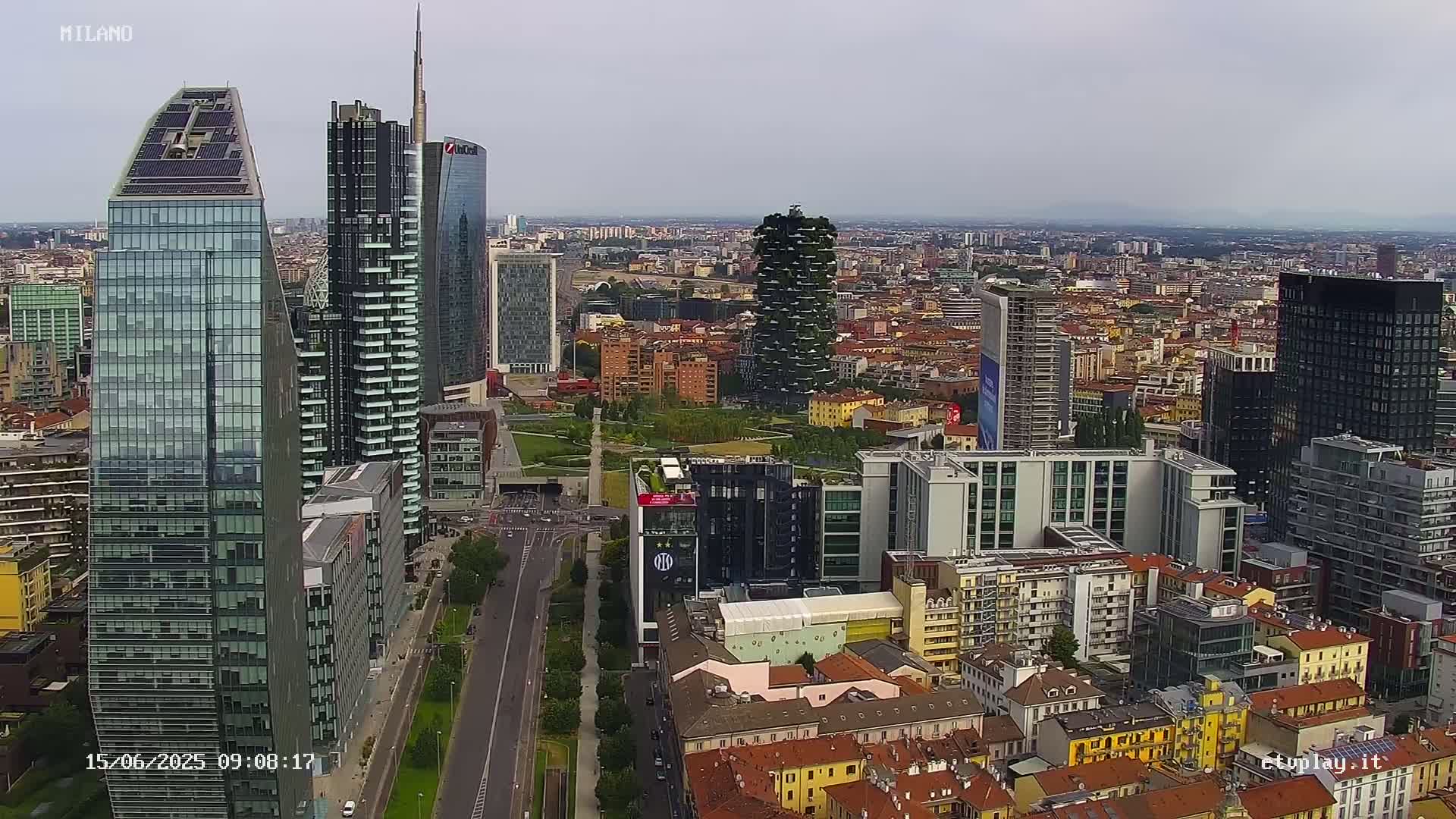 An aerial view of a city with many skyscrapers under an overcast sky shows a wide road leading through a park-like area between buildings.