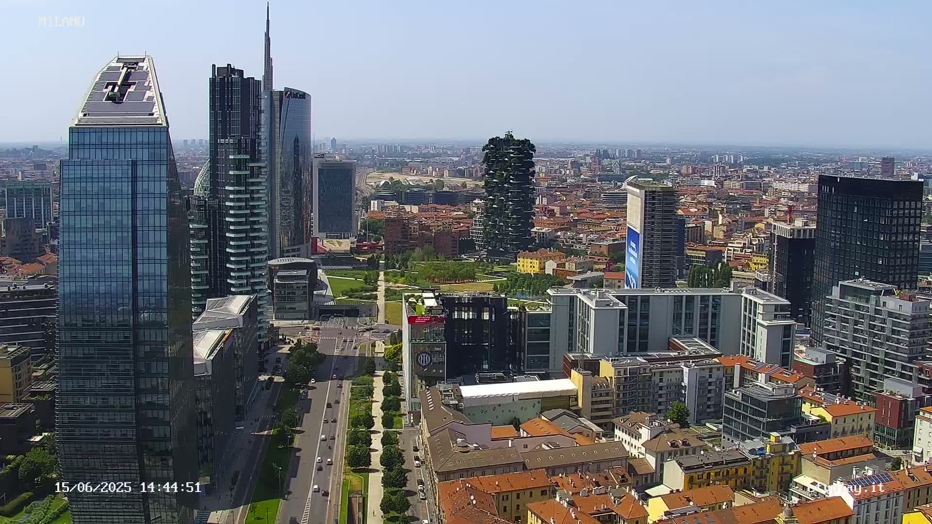 A high-angle, long shot on a sunny day shows a cityscape of modern skyscrapers and traditional low-rise buildings, with a park visible in the middle ground.