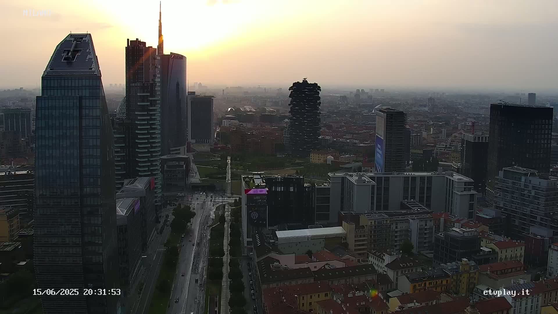A hazy sunset illuminates a cityscape of numerous skyscrapers and high-rise buildings, with a wide street and park visible below.