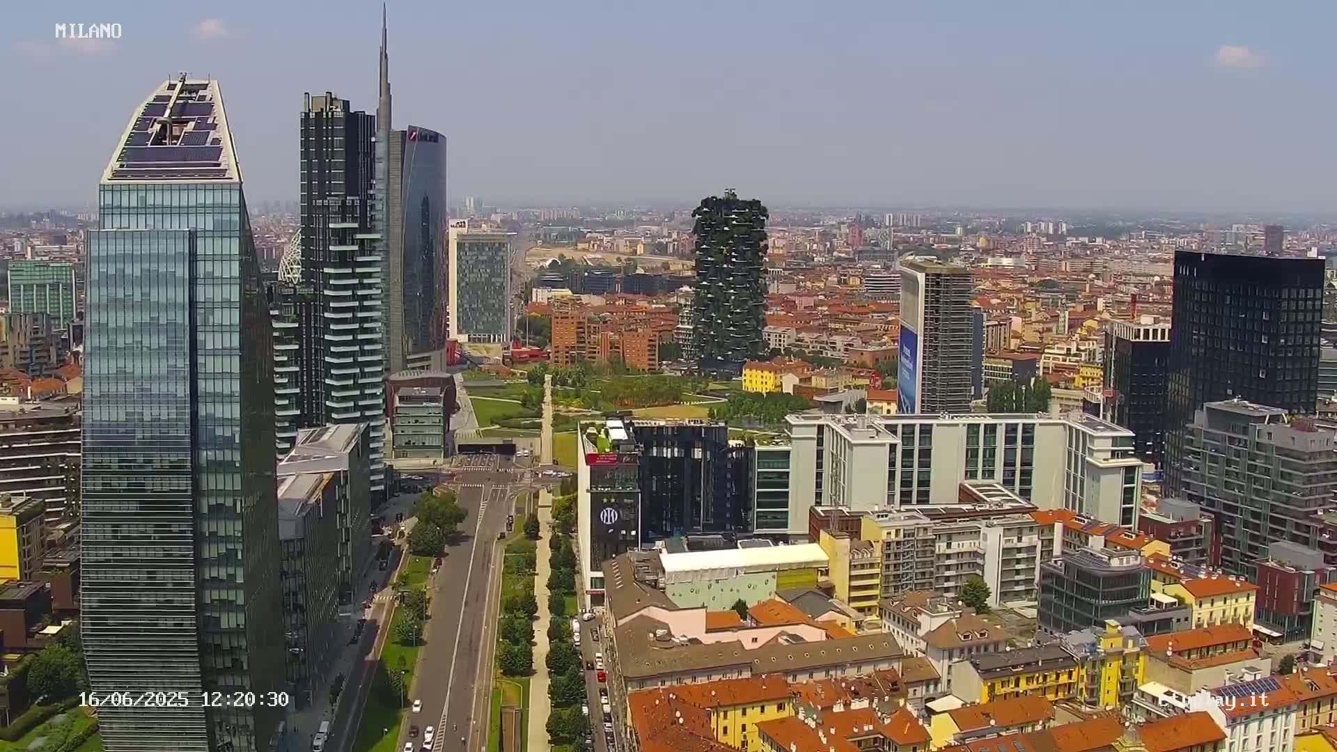 A high-angle, long shot on a sunny day shows a cityscape of many modern and traditional buildings, with a large road and green space in the foreground.