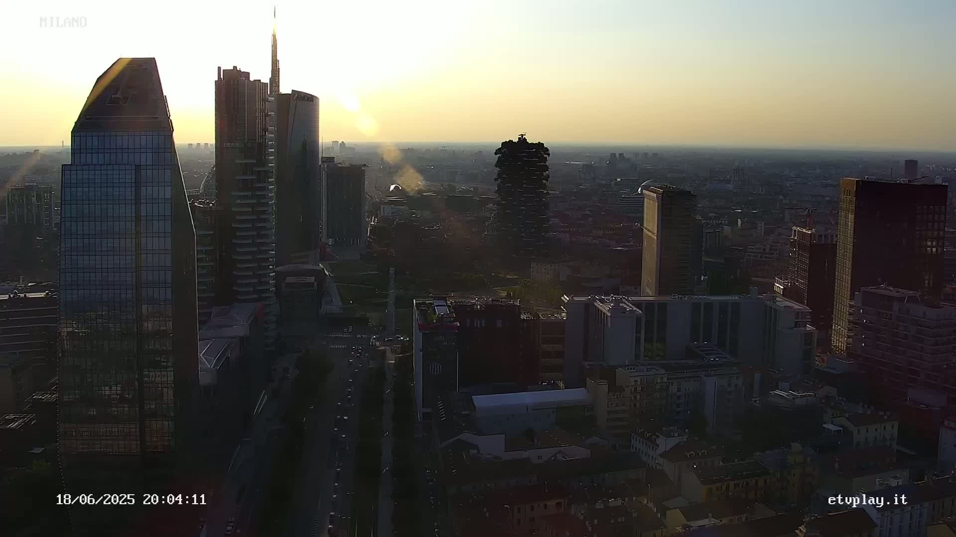An aerial view of a city skyline at sunset, with numerous skyscrapers silhouetted against a hazy, light orange sky.