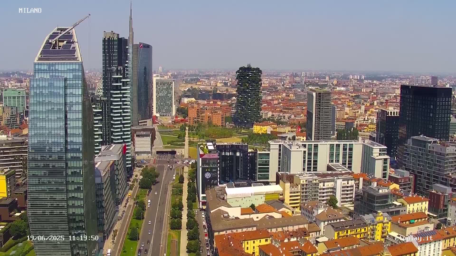 An aerial view shows a cityscape of modern and older buildings under a clear, sunny sky.
