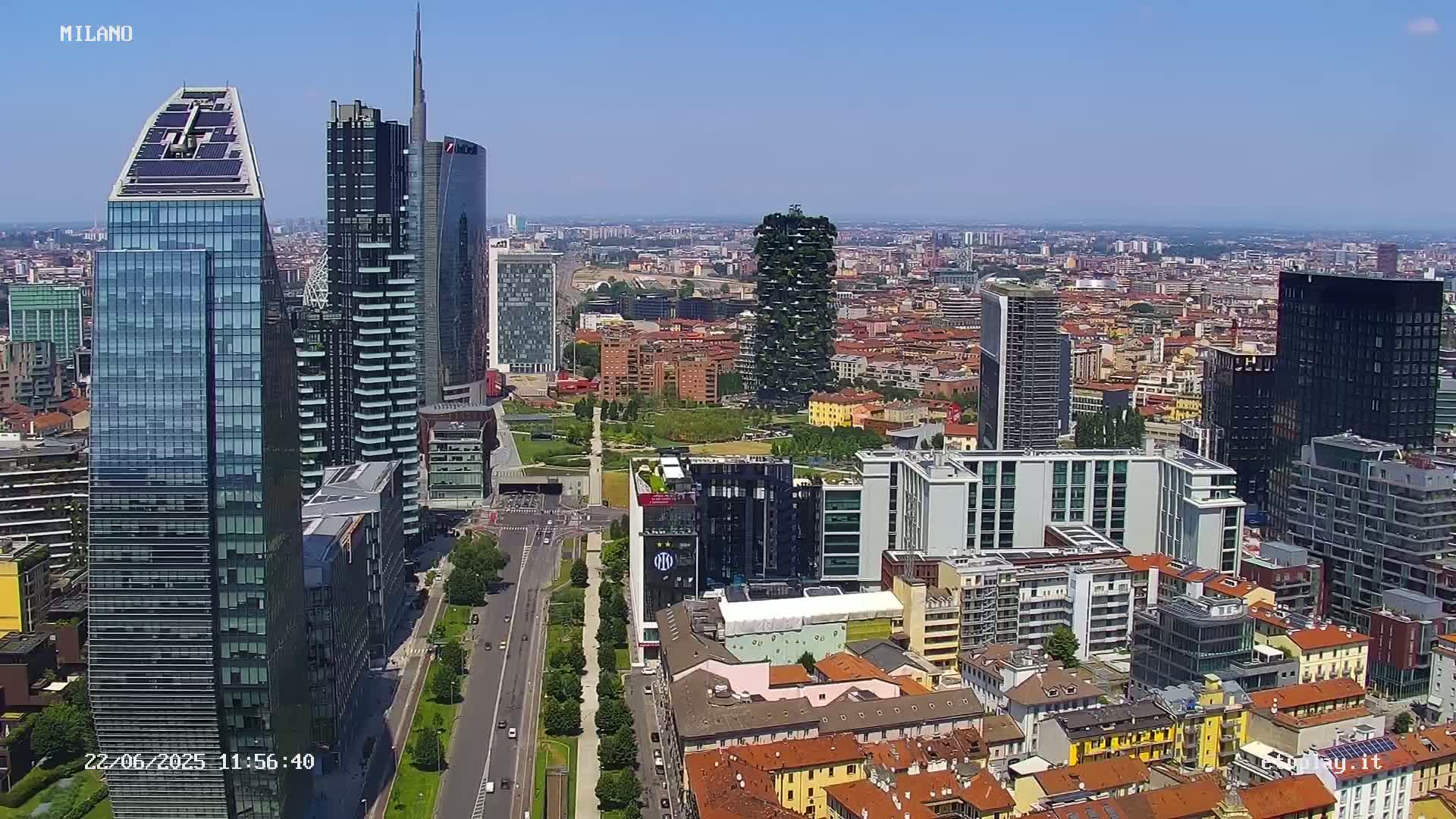 An aerial view of a city skyline on a clear, sunny day, showing numerous skyscrapers and surrounding buildings, with a wide road and green spaces visible.