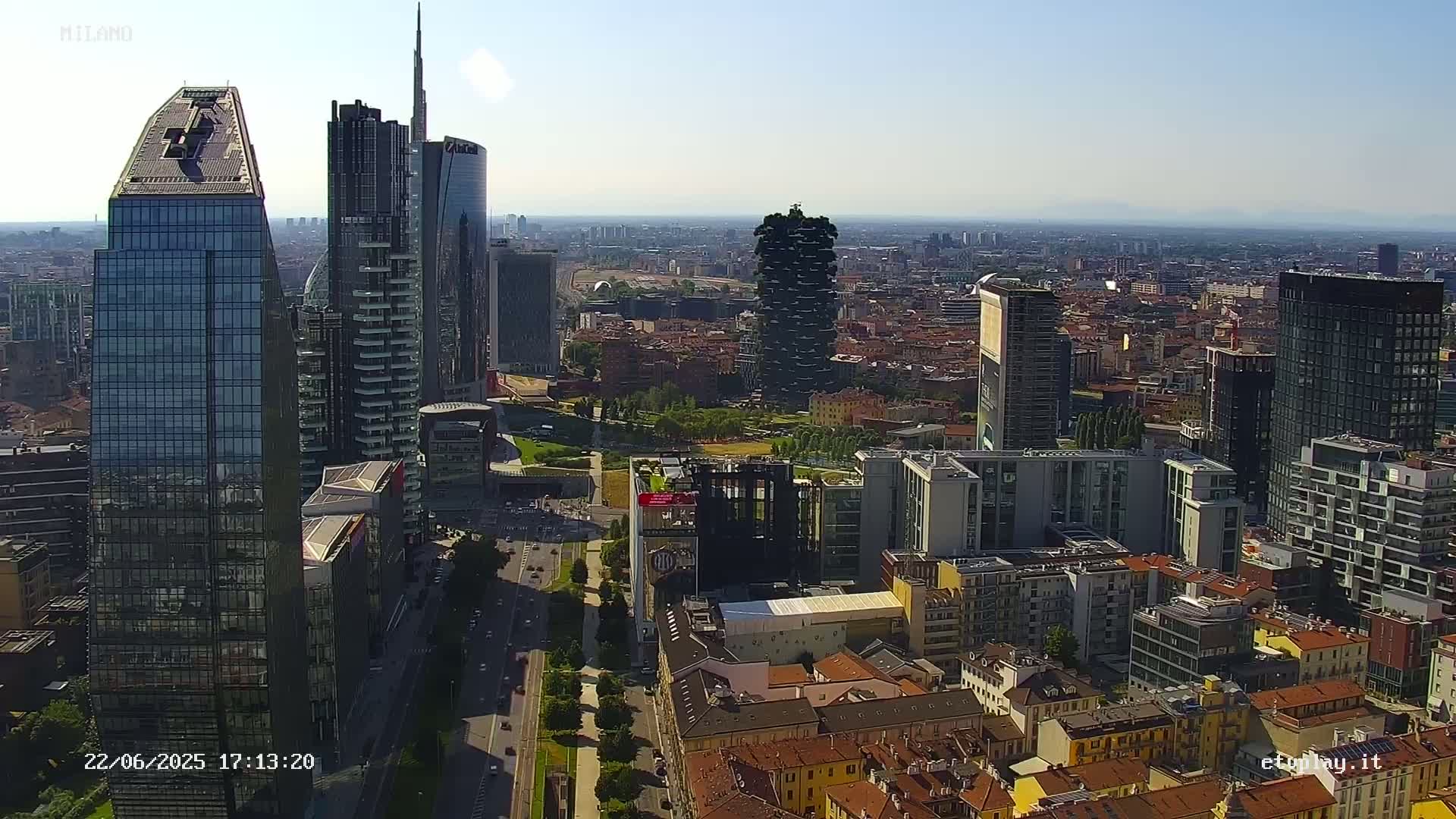 An aerial view of a city skyline on a clear, sunny day, showing numerous modern skyscrapers and surrounding low-rise buildings.