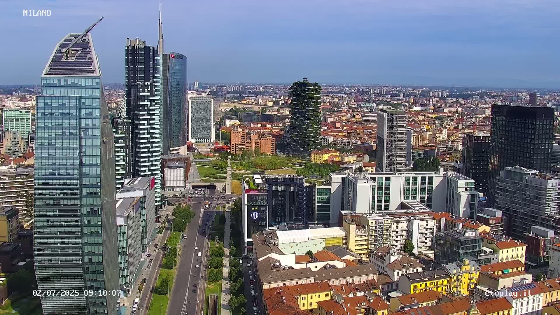 A high-angle, sunny view of a dense cityscape featuring numerous skyscrapers and a mix of modern and older buildings, with a large park visible in the mid-ground.
