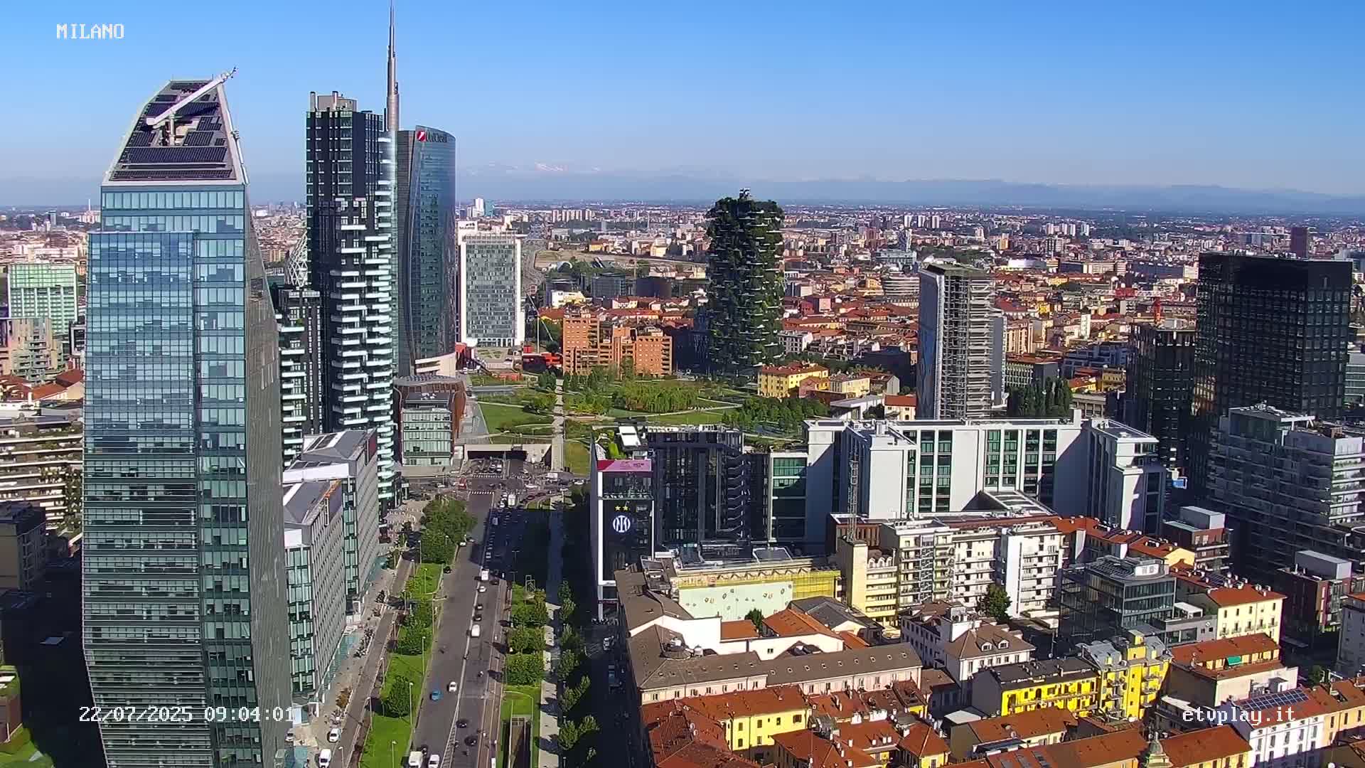 A high-angle, sunny-day view of a dense cityscape featuring numerous skyscrapers and a mix of modern and older buildings, with a park visible among the structures and mountains in the far distance.