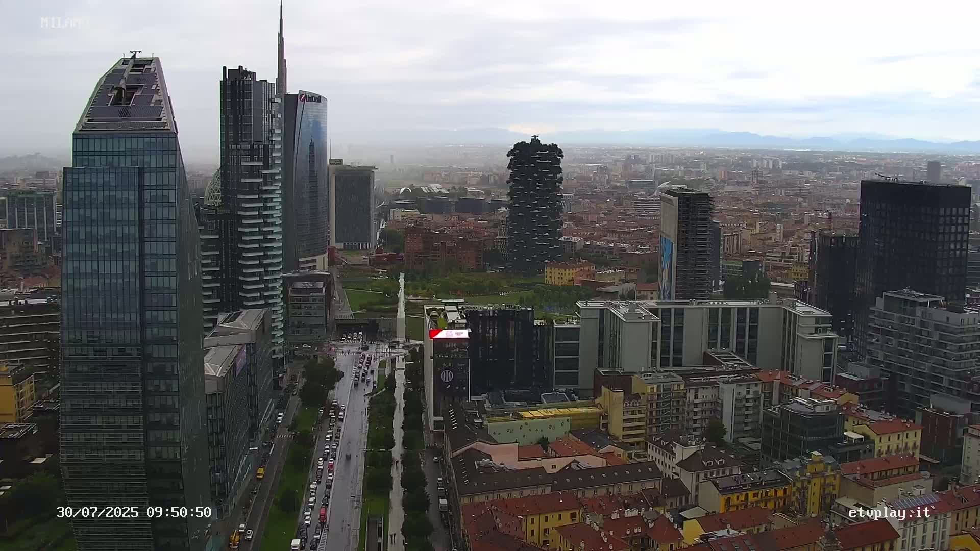 A high-angle, long shot of a city skyline on an overcast day, showing several modern skyscrapers and surrounding buildings along a wet street.
