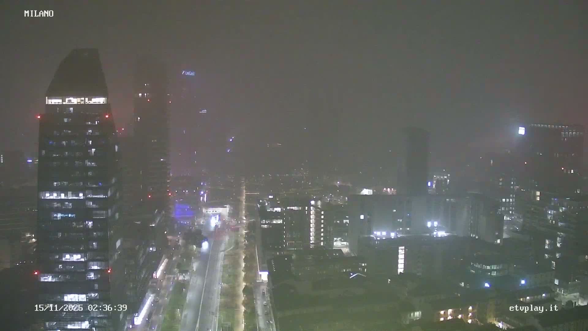 A hazy nighttime cityscape is visible through thick fog or mist, featuring illuminated skyscrapers and a bustling street with vehicle lights.