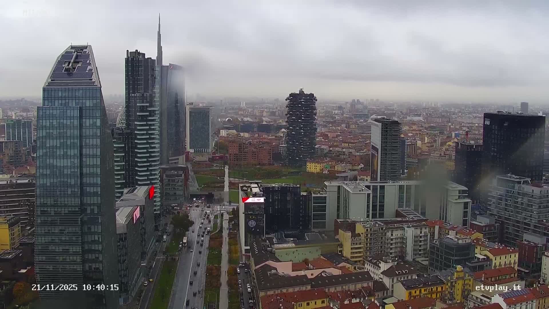 An expansive aerial view captures a bustling urban cityscape featuring striking modern skyscrapers, a busy wet street with traffic, and a blend of contemporary and traditional buildings, all under a heavily overcast and hazy sky.