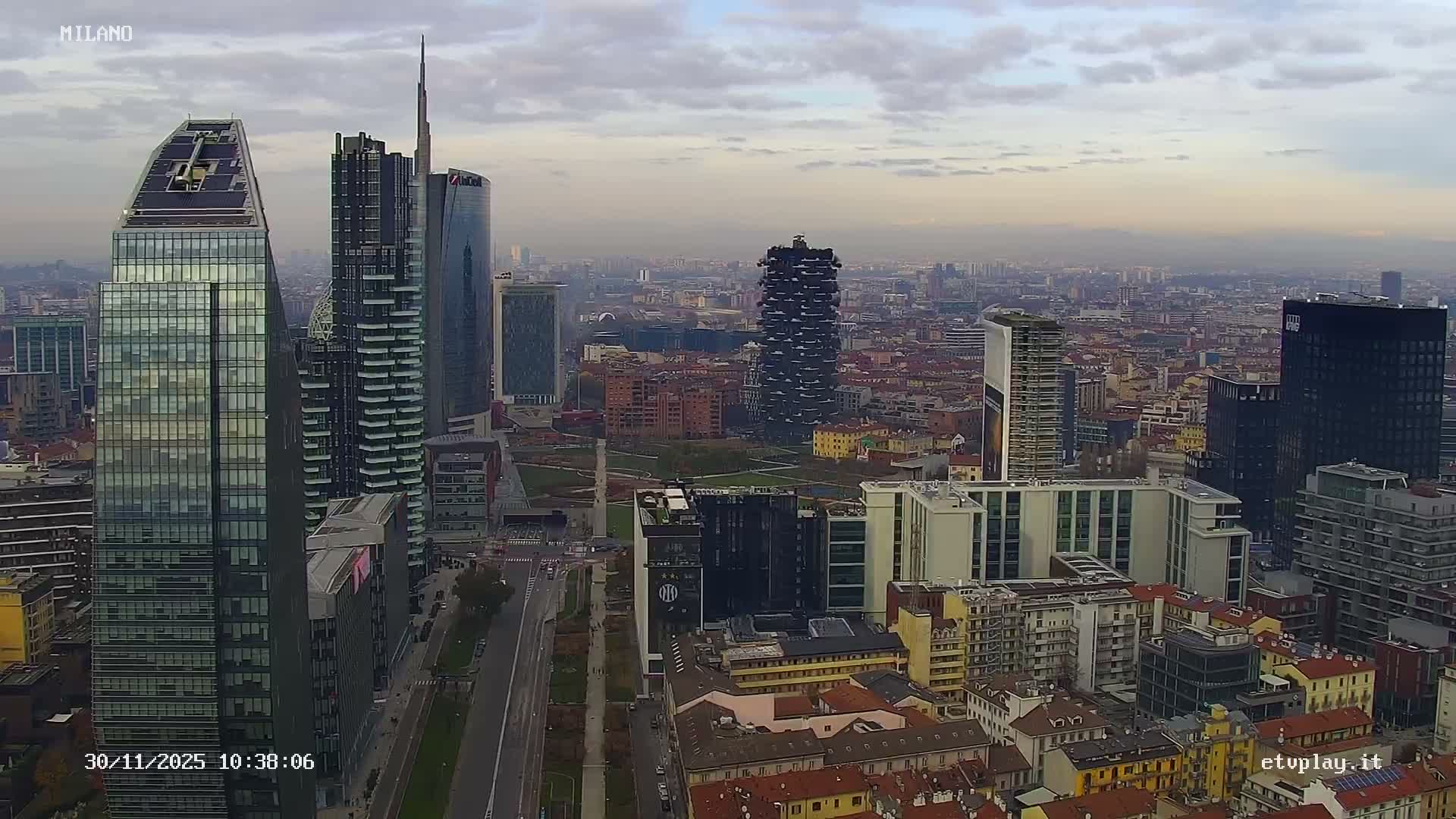 An overcast and hazy panoramic view of a modern city reveals a skyline dominated by sleek skyscrapers, including distinctive architectural structures and a tree-covered building, alongside a dense urban landscape of varied buildings and roads.
