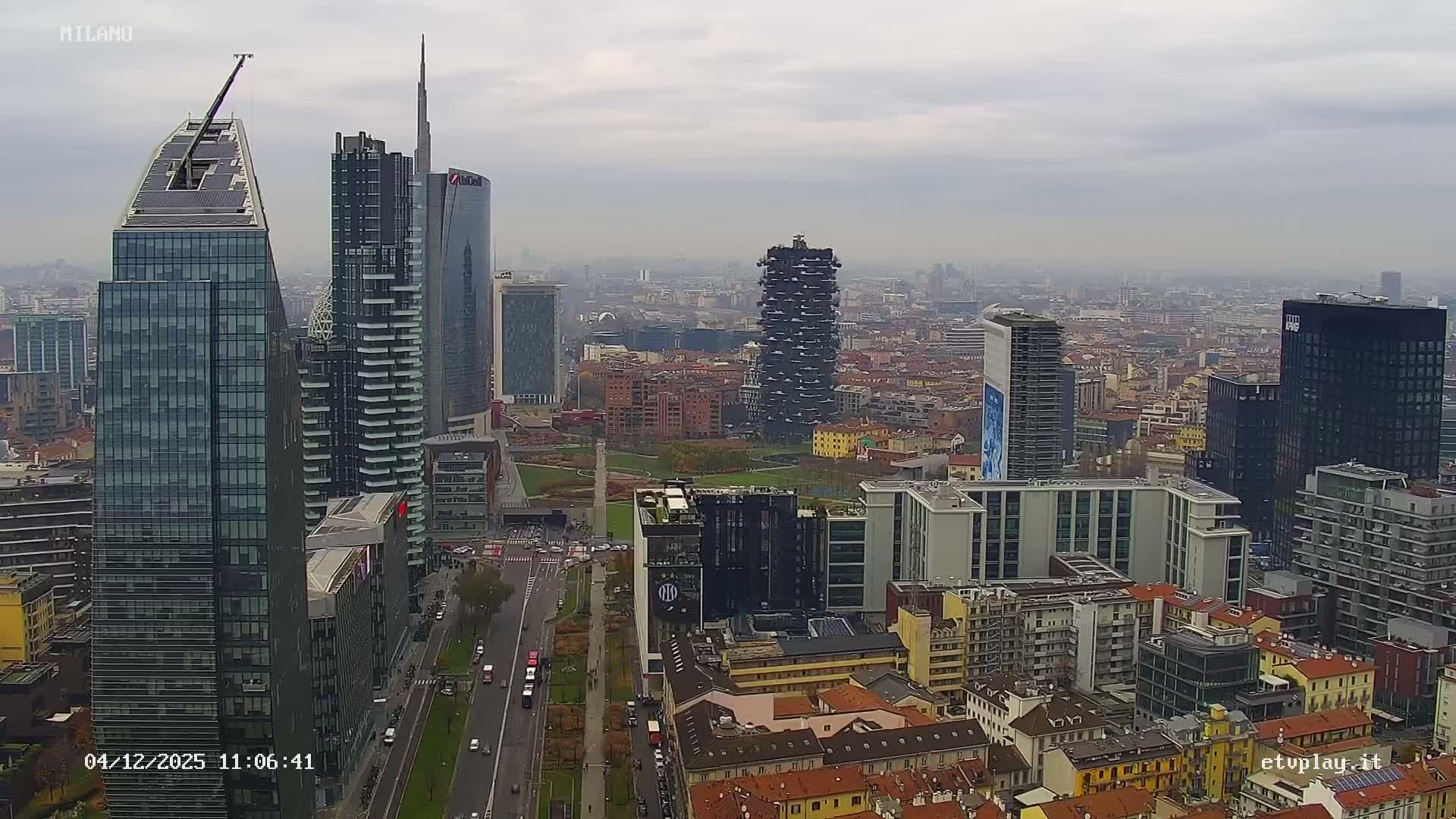 A sprawling cityscape of Milan, showcasing modern skyscrapers alongside traditional buildings and a central park with active roads, is viewed under an overcast and hazy sky.