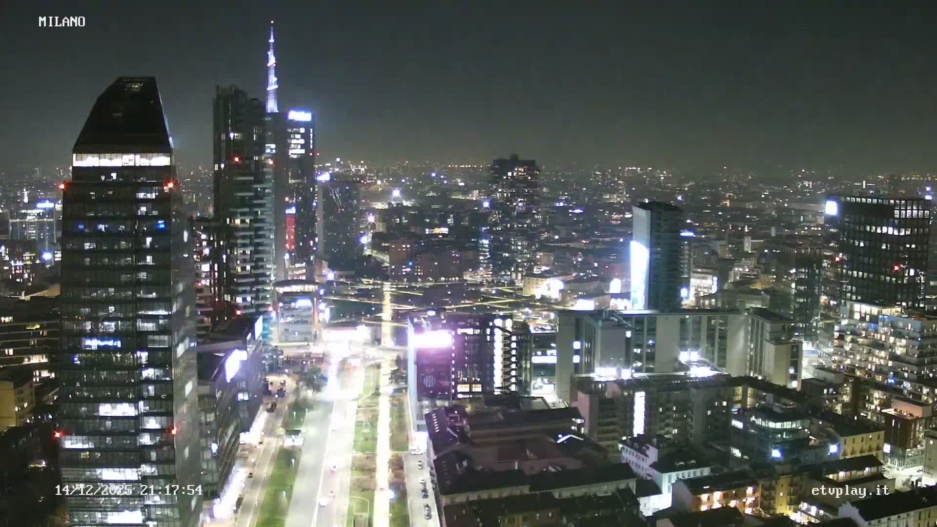 A sprawling cityscape of Milan, showcasing modern skyscrapers alongside traditional buildings and a central park with active roads, is viewed under an overcast and hazy sky.