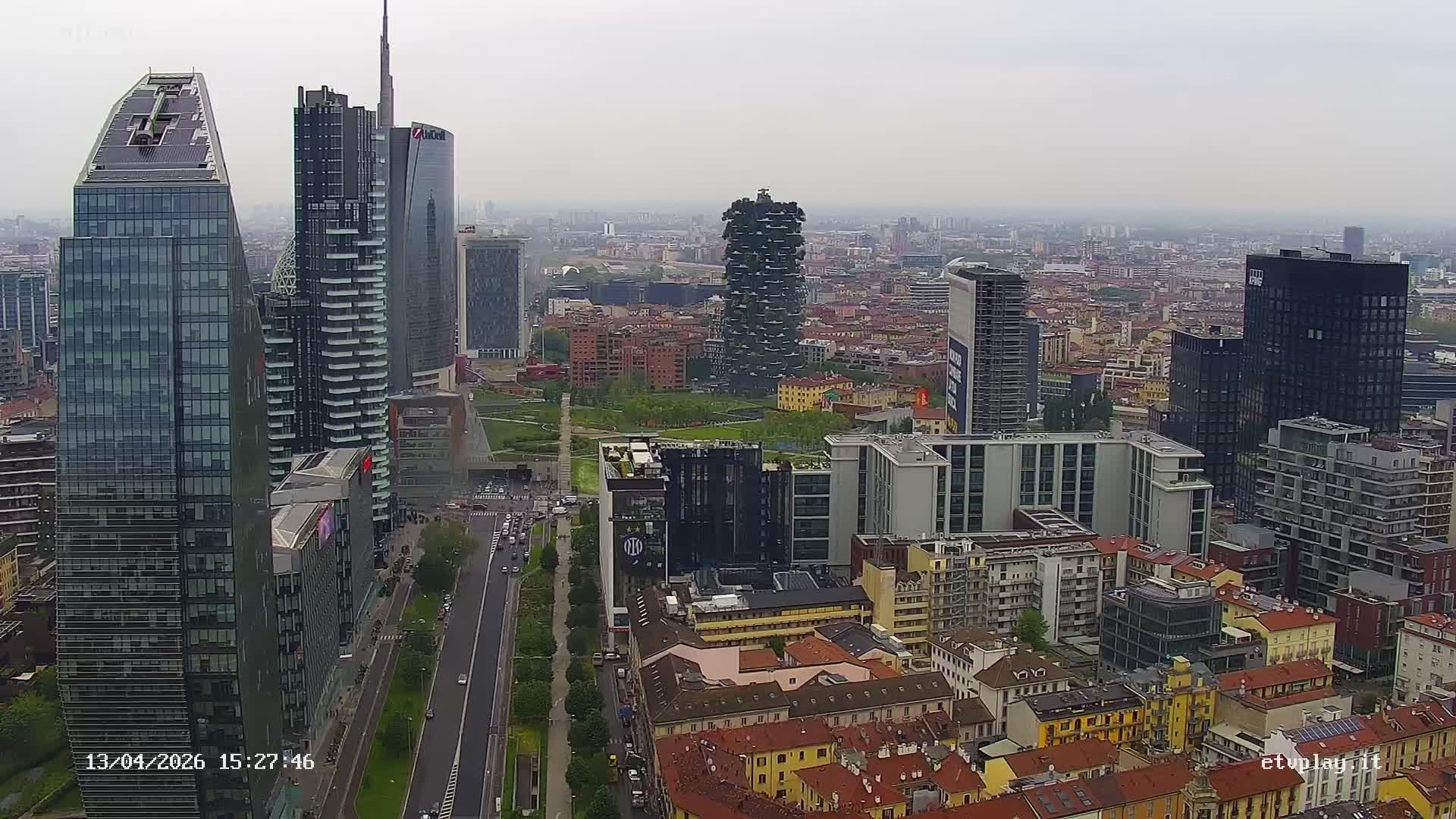 A sprawling cityscape of Milan, showcasing modern skyscrapers alongside traditional buildings and a central park with active roads, is viewed under an overcast and hazy sky.