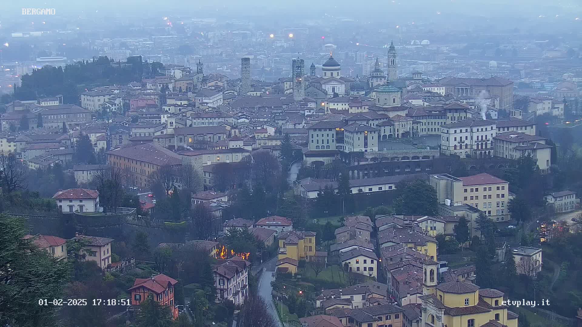 A sprawling city, possibly Bergamo, is viewed from above on a hazy or foggy day.