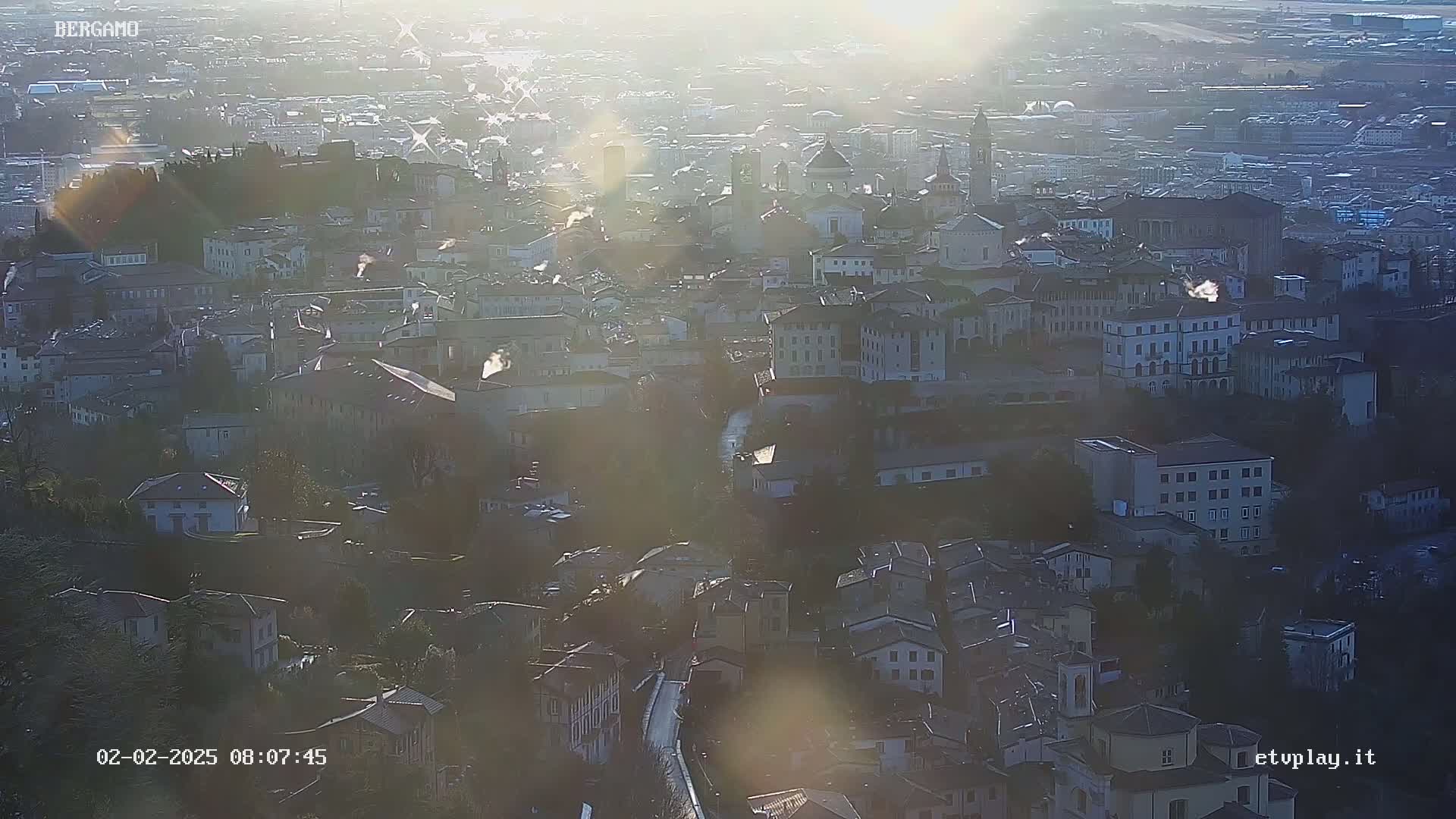 A hazy sun shines on a sprawling city of closely packed buildings, viewed from a high vantage point.