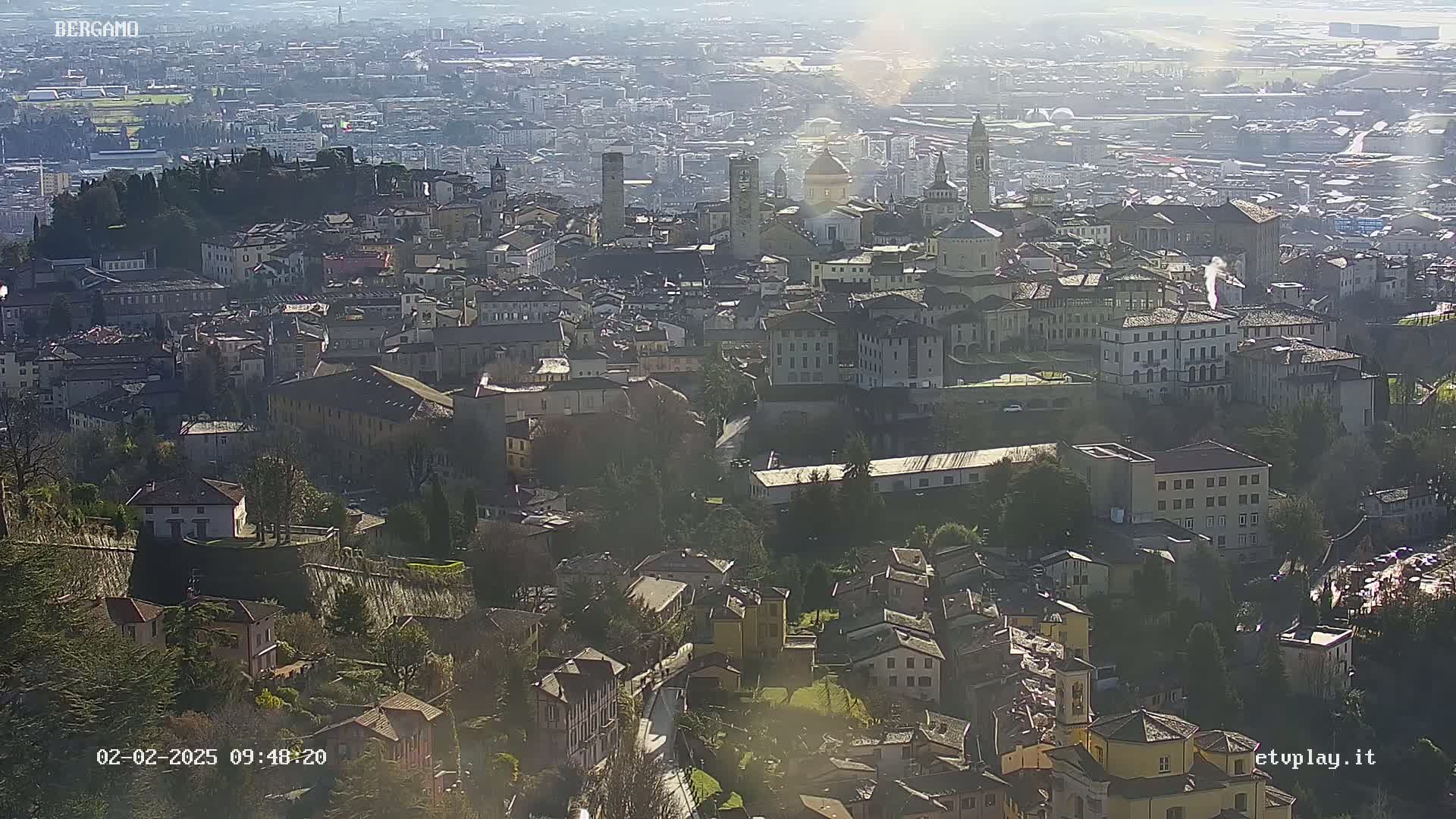 A hazy sun shines on a sprawling city of closely packed buildings, many light-colored, nestled in a valley.