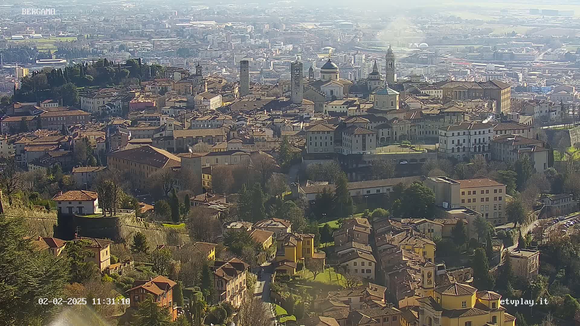 A sprawling city of tan and brown buildings sits nestled in a valley on a sunny day with hazy skies.