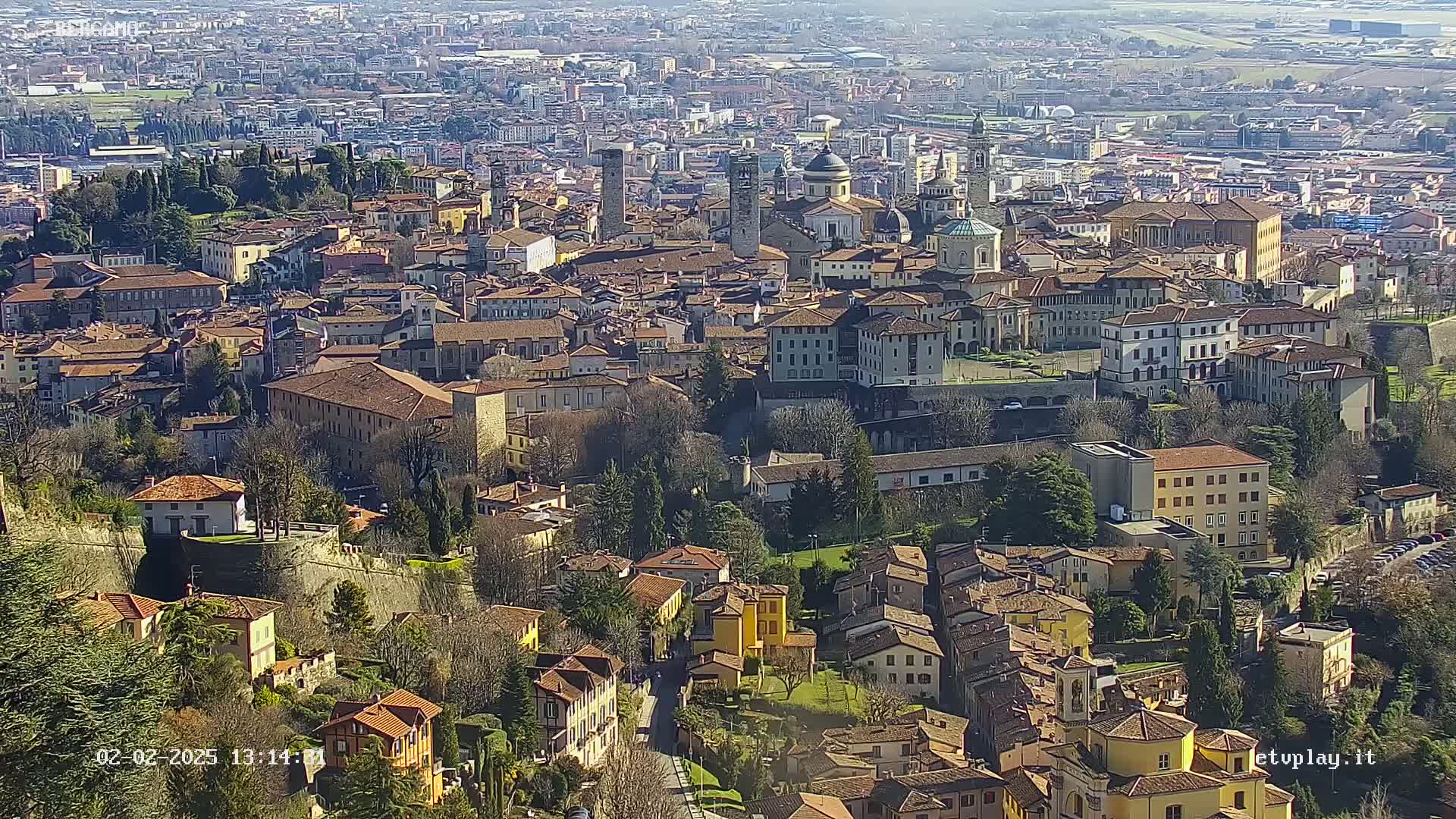 A sprawling city of terracotta-roofed buildings, nestled within ancient stone walls, is visible under a clear, sunny sky.