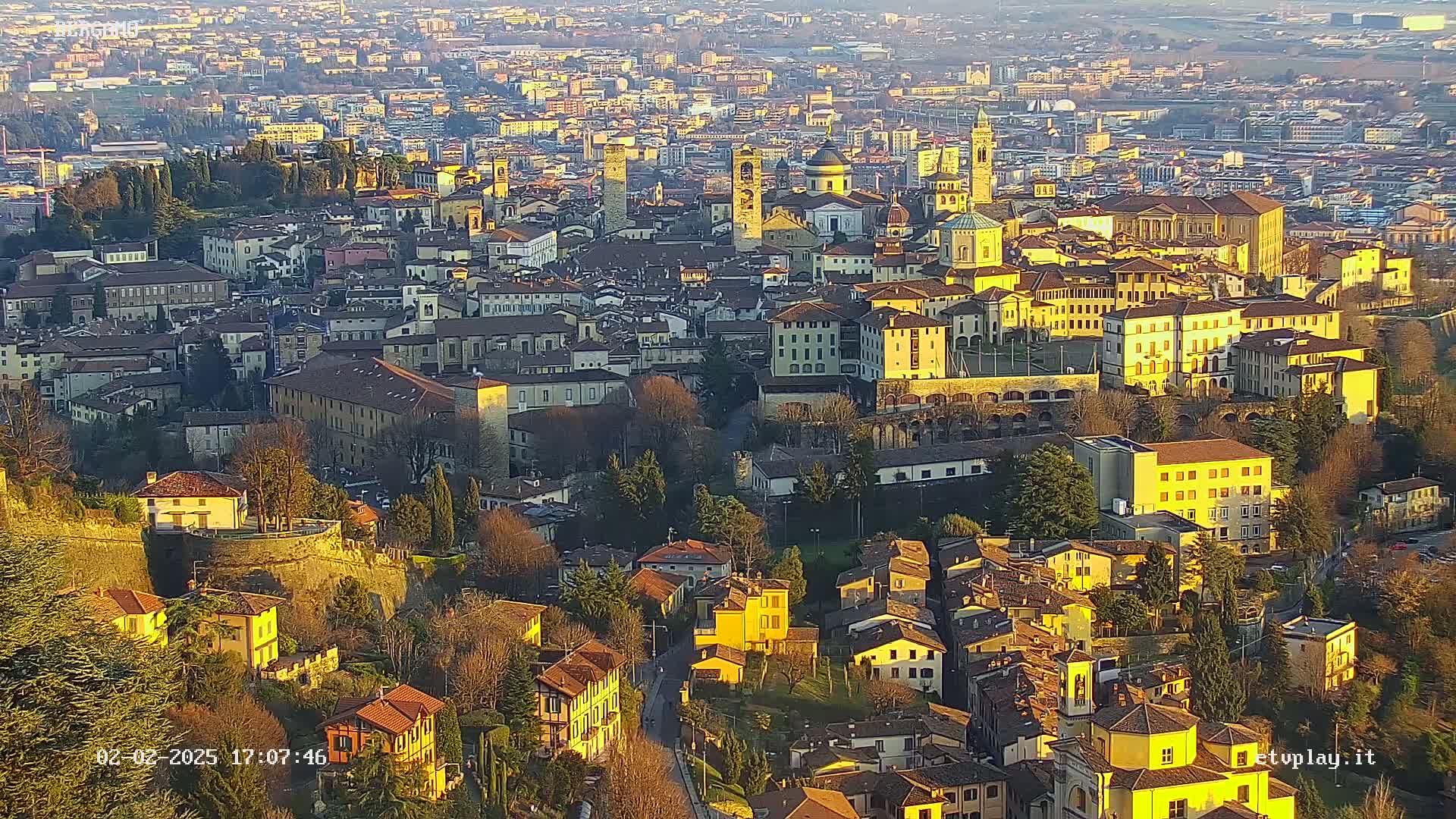 An aerial view of a city, bathed in the golden light of late afternoon or early evening, shows numerous buildings of varying heights and styles nestled together on a hillside.
