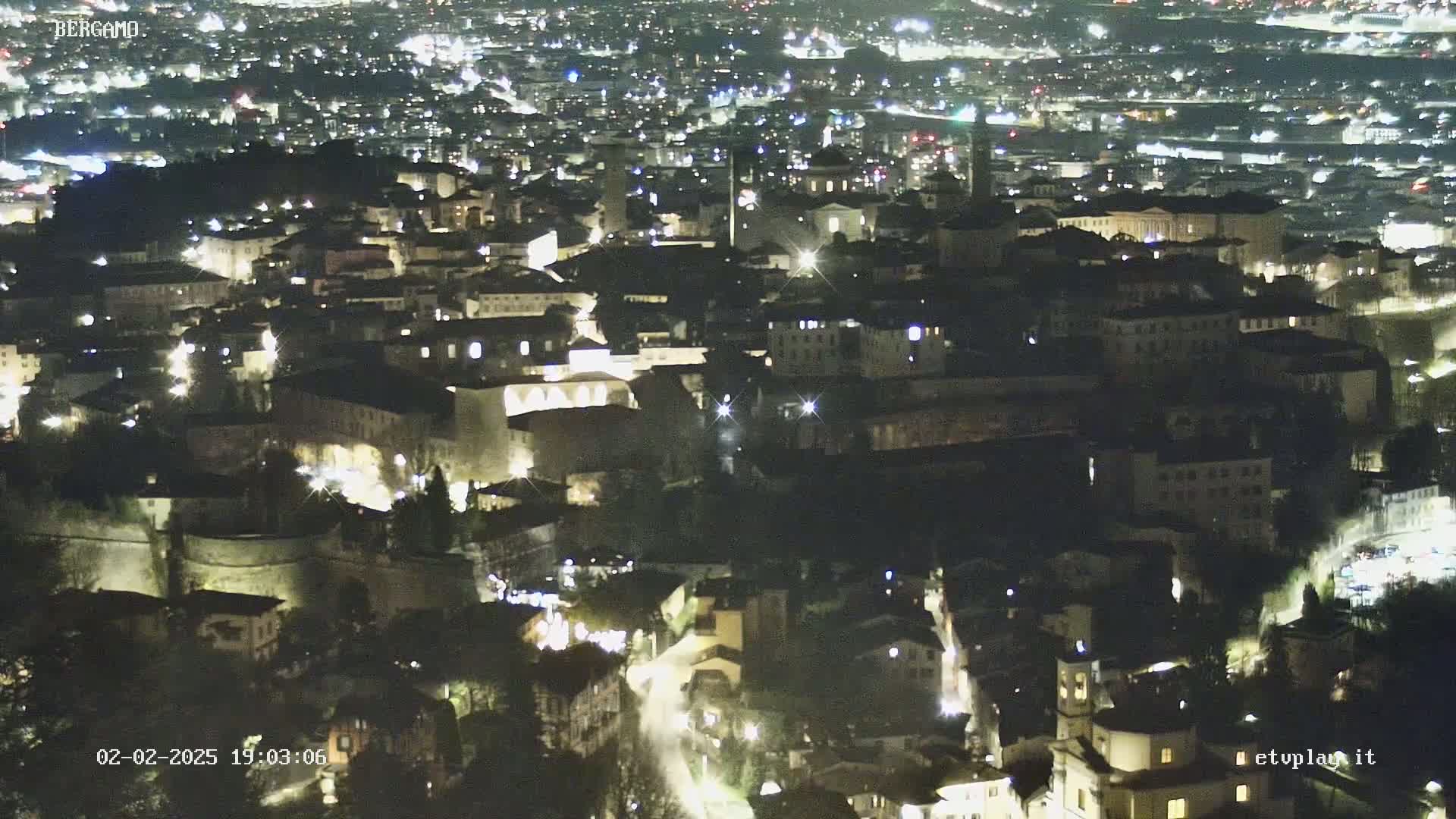 A nighttime aerial view of a city with numerous illuminated buildings, nestled within surrounding hills.