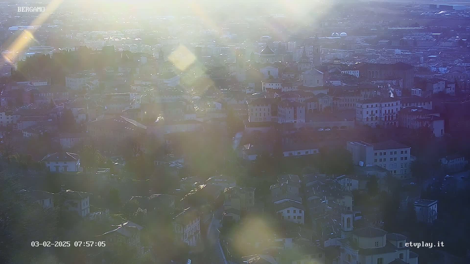A hazy, sunlit, high-angle view of a dense cityscape, possibly in the early morning or late afternoon.