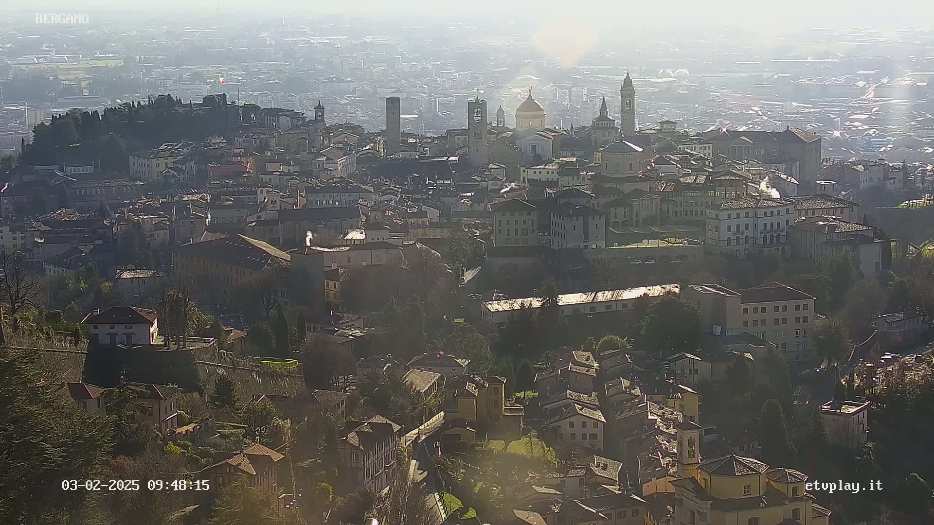 A hazy, sunny day overlooks a sprawling city with numerous buildings, including churches and towers, nestled amongst hills and greenery.