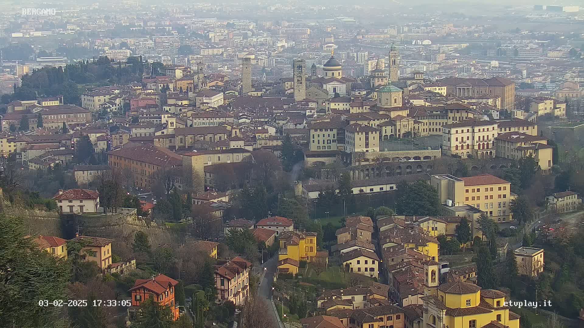 A hazy, high-angle view of a dense, historic city with many tan and brown buildings, including several churches and a large stone wall, nestled within a landscape of green trees.