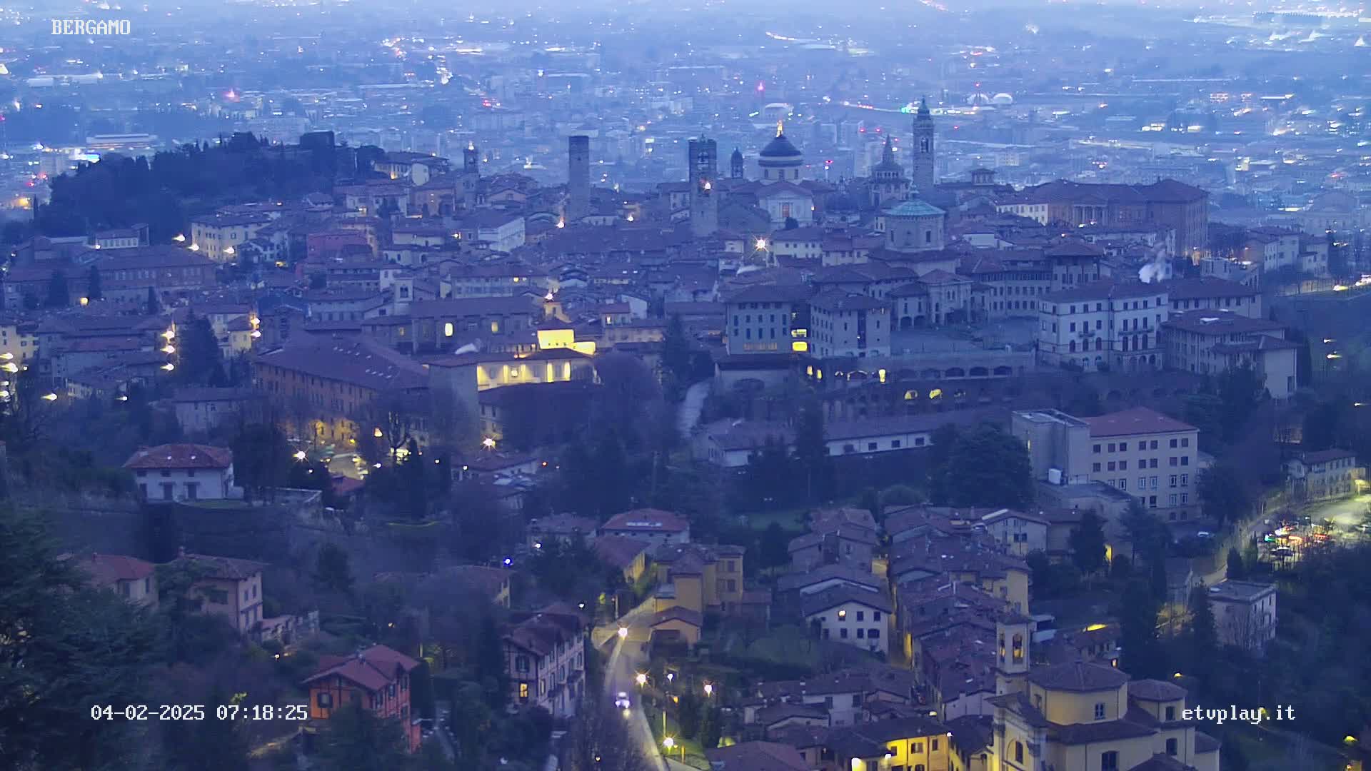 A nighttime, aerial view of a city with numerous buildings illuminated, nestled in a valley under a twilight sky.