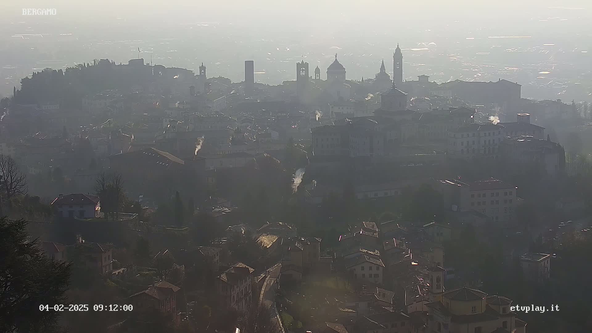 A hazy, distant view of a city with numerous buildings and several prominent towers and domes, under a mostly clear sky.