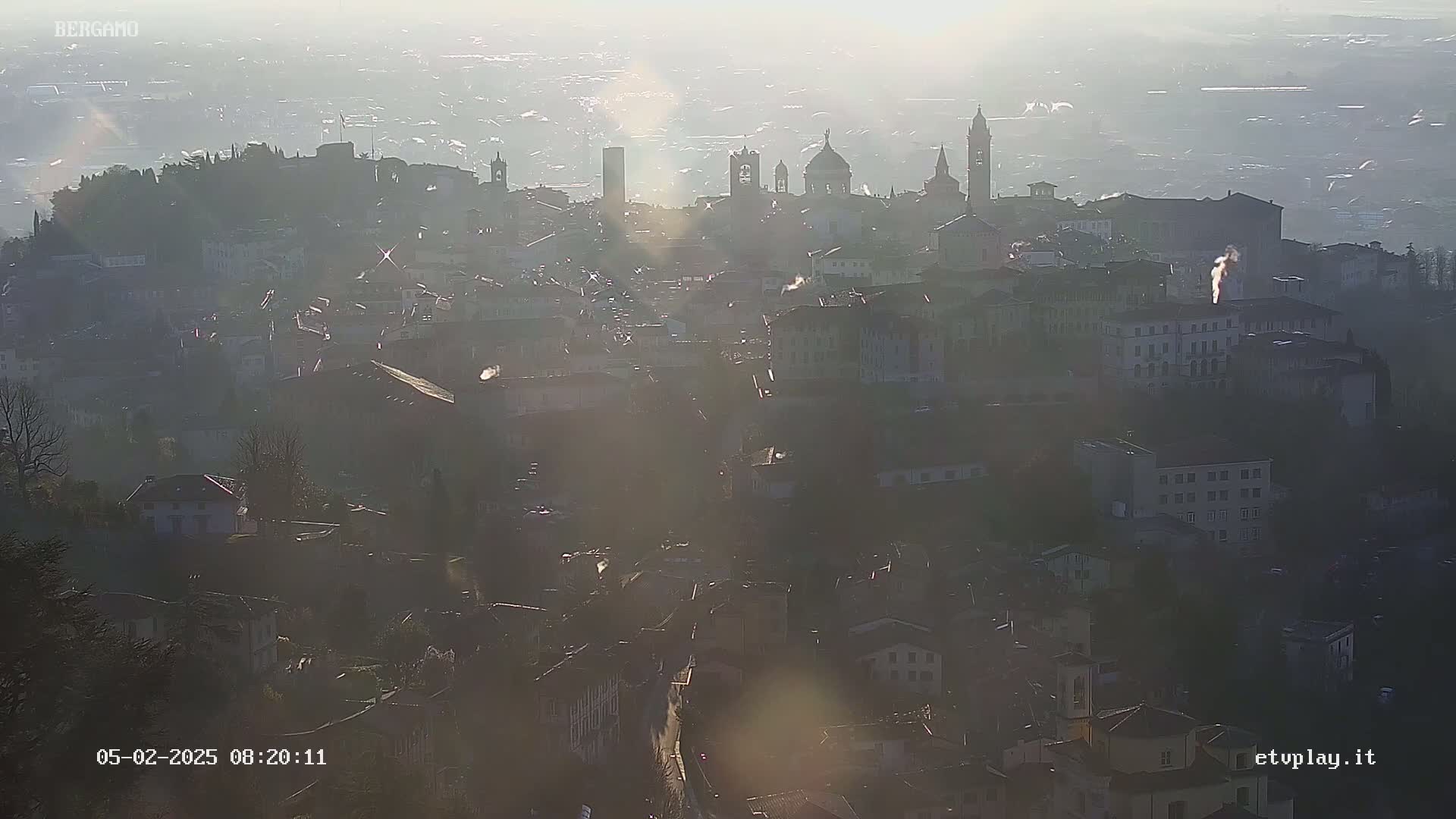 A hazy, sunlit view of a city with numerous buildings and several prominent structures, possibly churches or towers, is visible from a high vantage point.