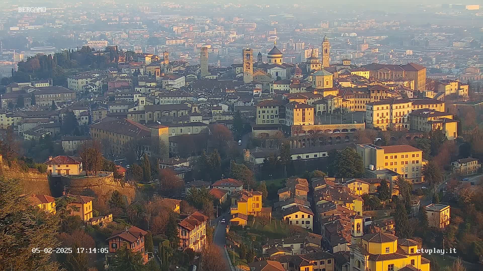 A hazy, sunlit view of a dense collection of buildings, situated on a hill, with a surrounding cityscape visible in the distance.