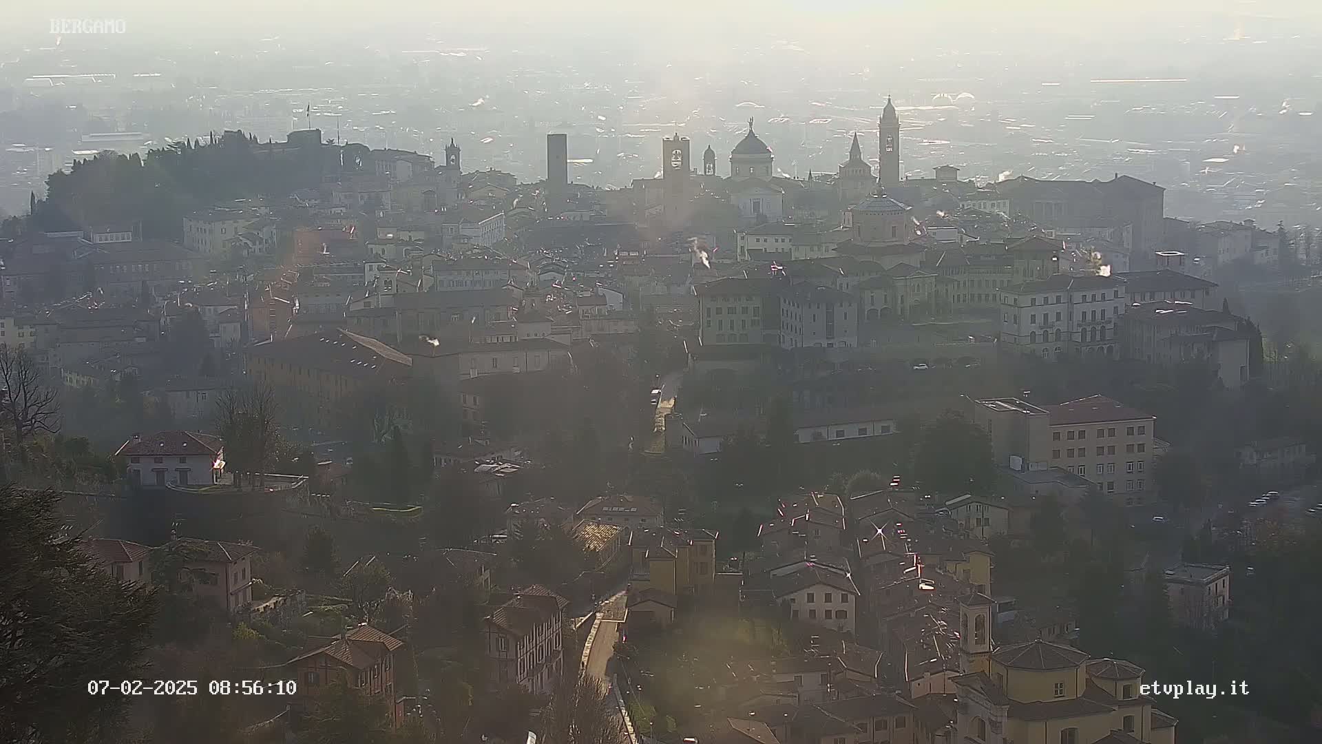 A hazy, sunlit view of a sprawling city with numerous buildings, including several churches and towers, nestled among hills.