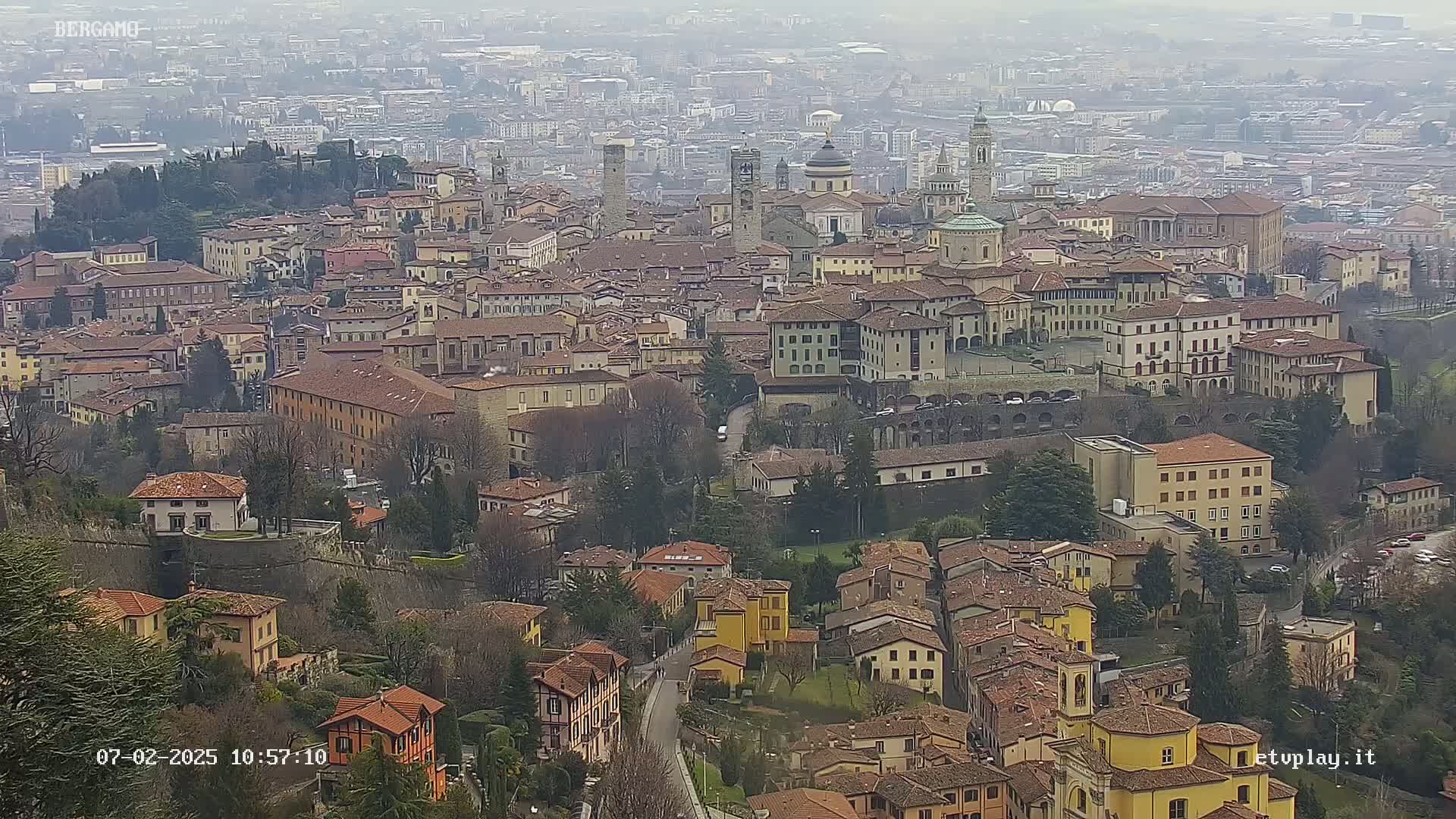 A sprawling city of many low-rise buildings with red-tiled roofs, interspersed with taller structures and churches, is viewed from above on an overcast day.
