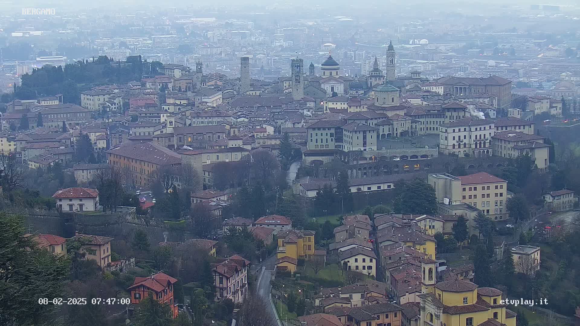 A hazy, overcast view of a densely packed European city with numerous buildings, churches, and a river running through it.