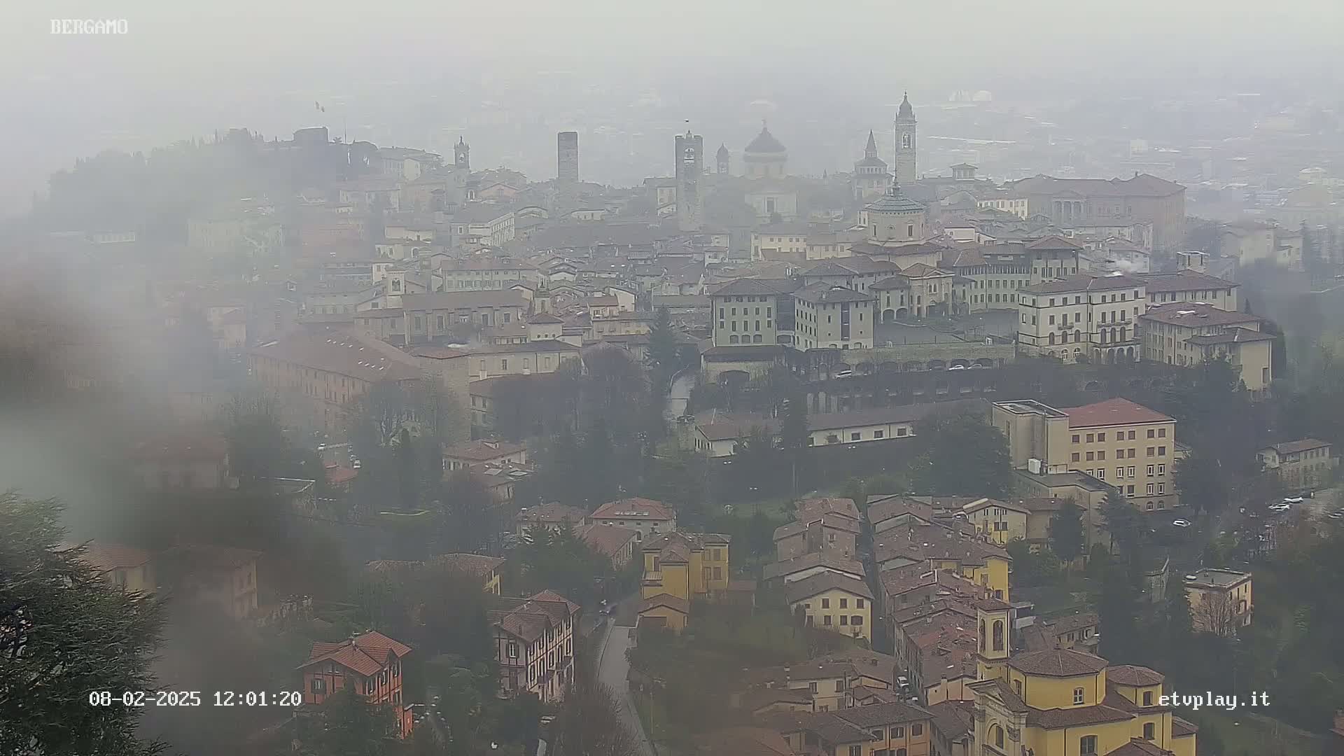 A misty, overcast view of a sprawling city with numerous buildings, including several prominent structures that appear to be churches or towers.