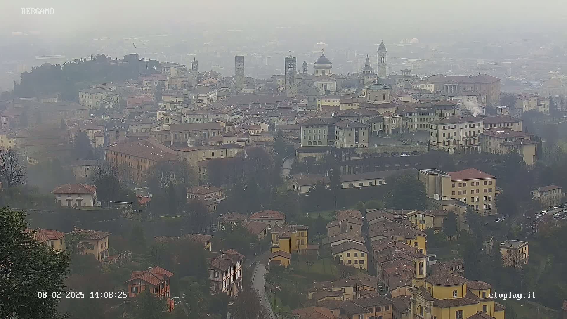 A hazy view of a European city with many closely packed buildings, showing a mix of architectural styles and a river or stream running through the lower part of the scene.