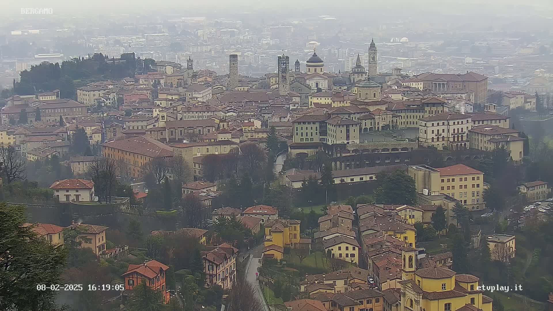 A hazy view of a city with numerous buildings, including churches and towers, nestled among trees and surrounded by a sprawling landscape.