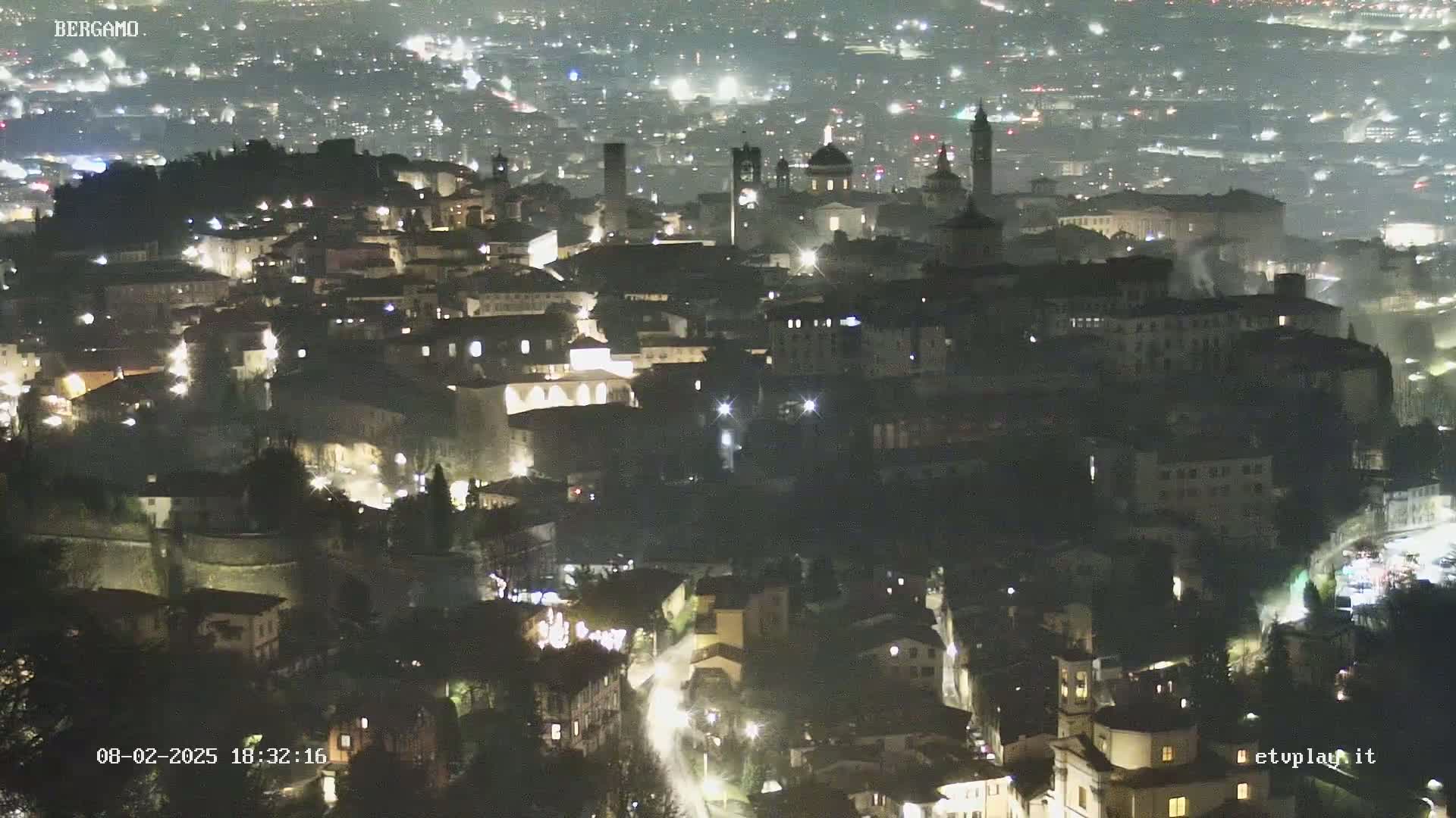 A nighttime, aerial view of a city with many illuminated buildings, including several tall structures that appear to be churches or towers.