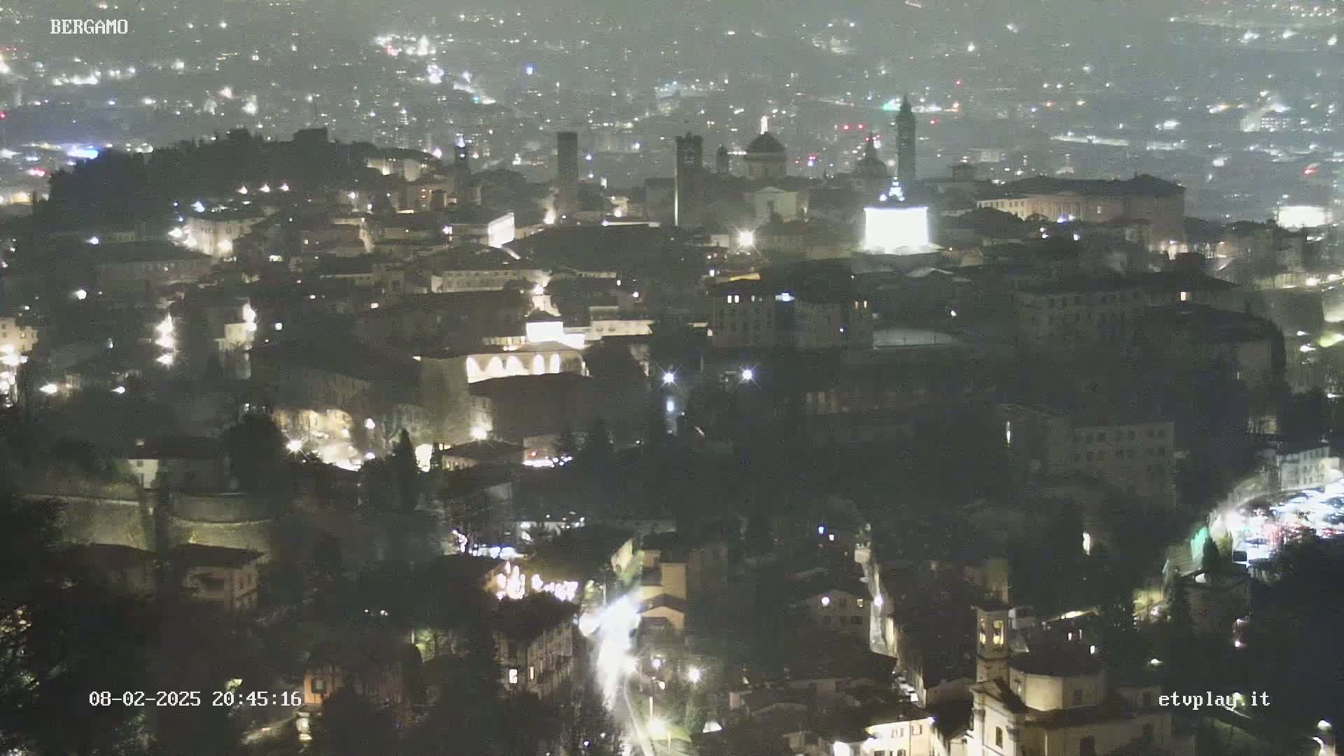A nighttime, aerial view of a city with faint light rain or mist, showing a densely packed collection of buildings illuminated by streetlights.