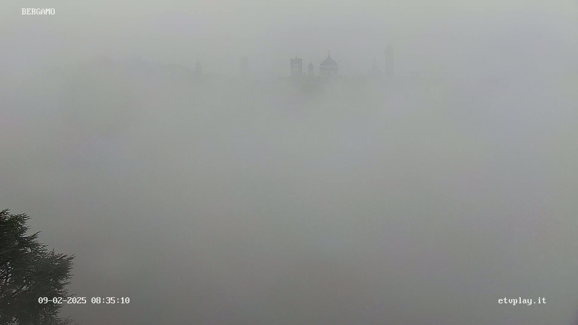 A cityscape, mostly obscured by a thick fog, shows the faint silhouettes of buildings, including what appears to be a church or cathedral.