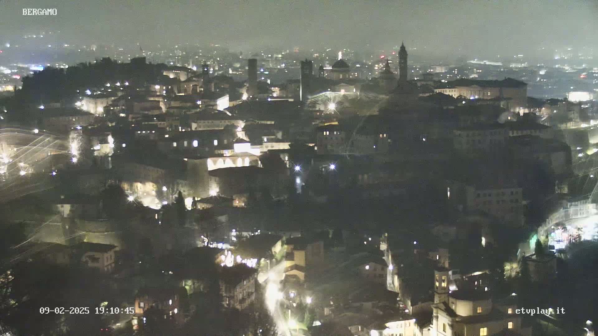 A nighttime, hazy view of a city with illuminated buildings and streets, possibly with fireworks in the distance.