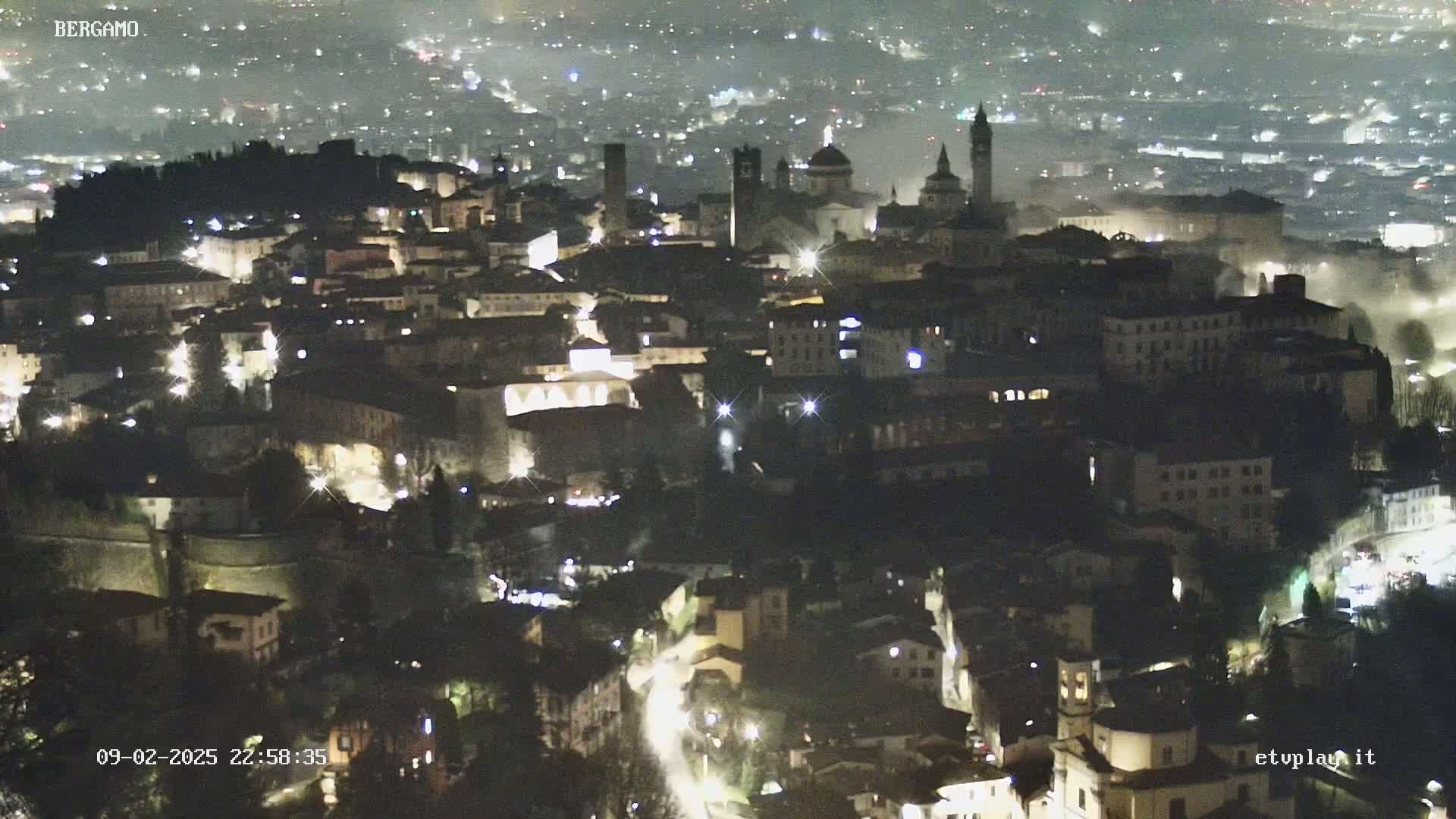 A nighttime aerial view of a city illuminated by lights, with a hazy atmosphere.