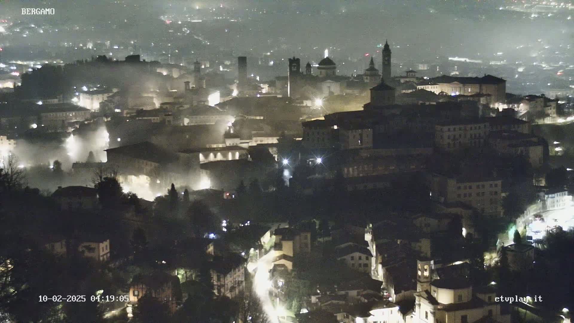 A nighttime view of a city shrouded in fog, with dimly lit buildings and streets visible.