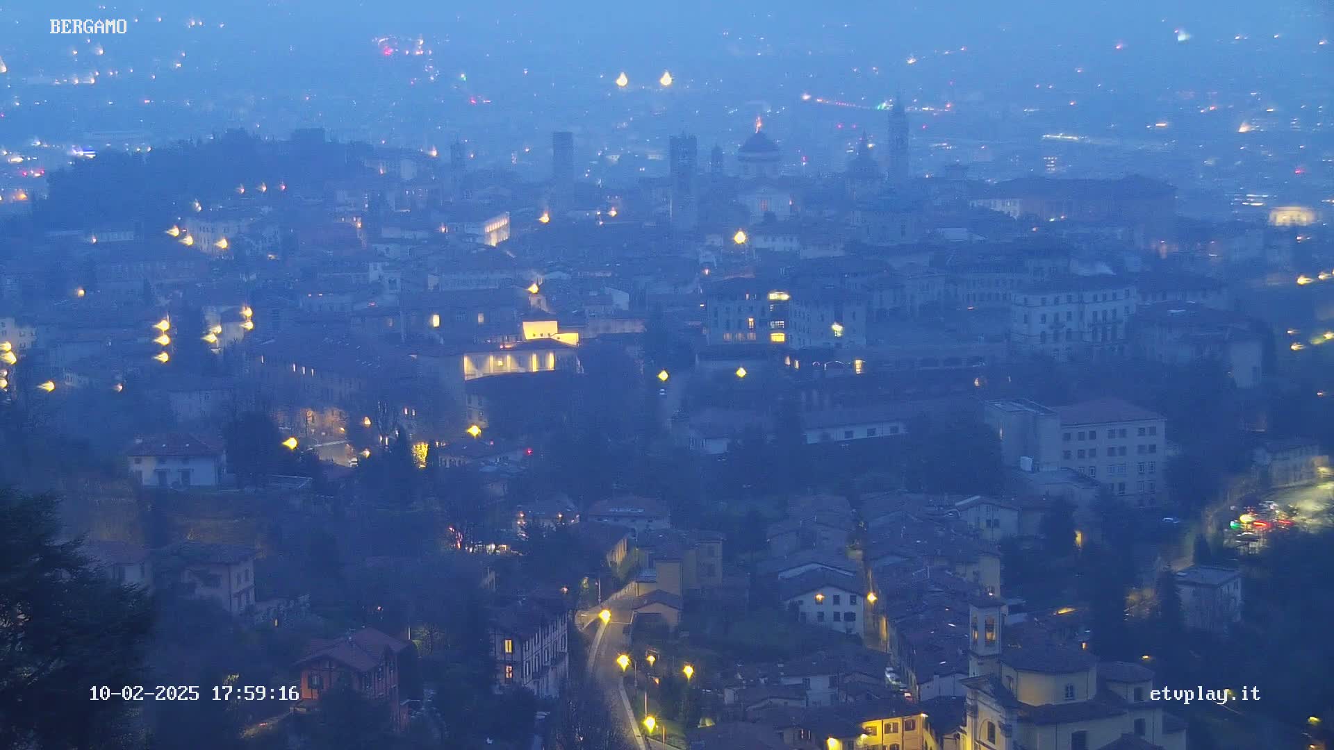 A hazy, twilight view of a city at night, showing numerous buildings with illuminated windows and streetlights.
